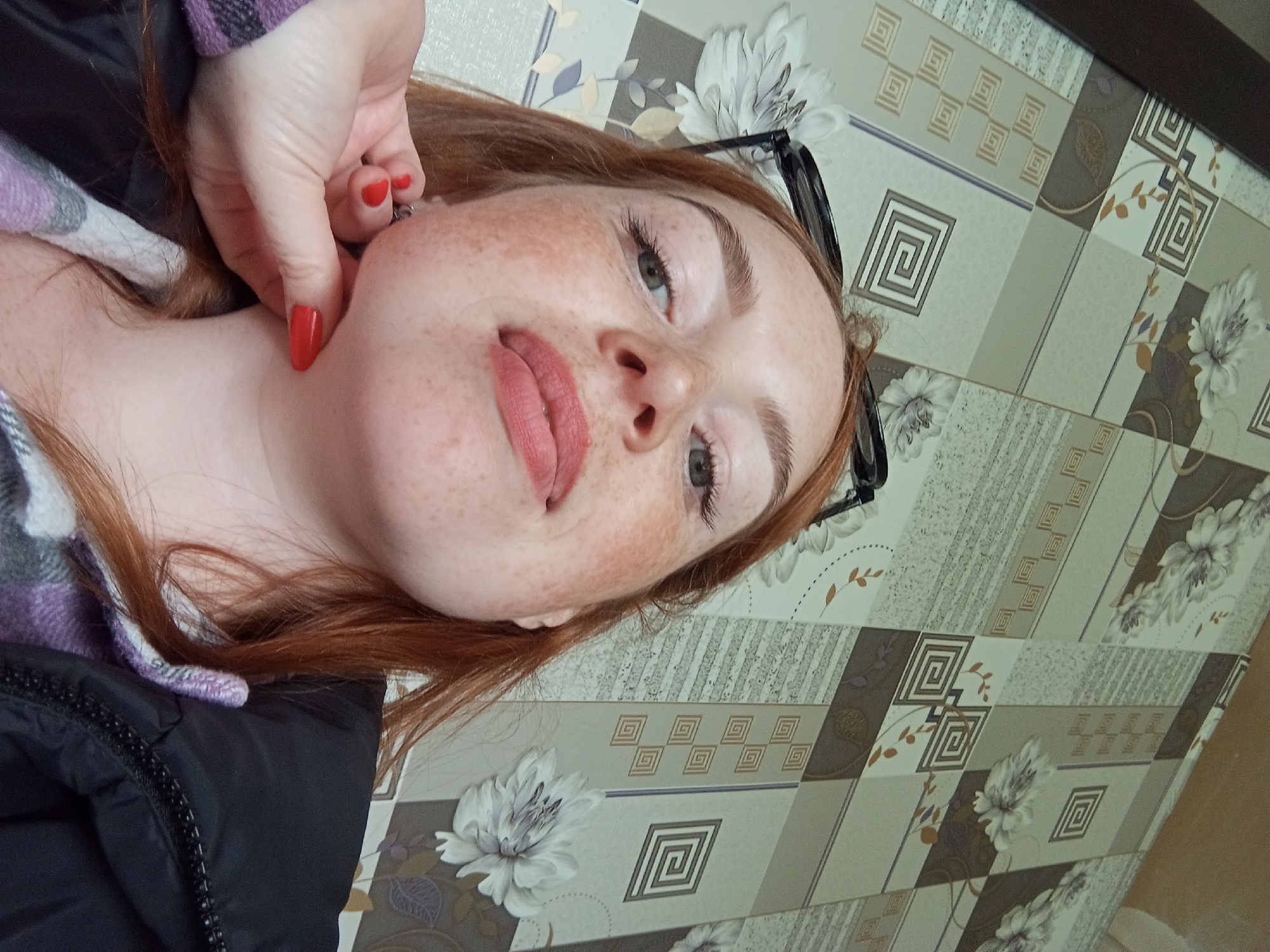 A close-up, slightly angled portrait depicts a fair-skinned individual with red hair and numerous freckles. The person is partially visible, with their face, neck, and a section of their upper body in view. A hand with red nail polish is touching the person's face near the cheek. A pair of black glasses are positioned on the right side of the frame, near the person's hair. The individual is wearing what appears to be a black jacket and a patterned shirt. The background consists of a surface covered with a geometric and floral pattern. The location is identified as Yurgamysh, Russia.