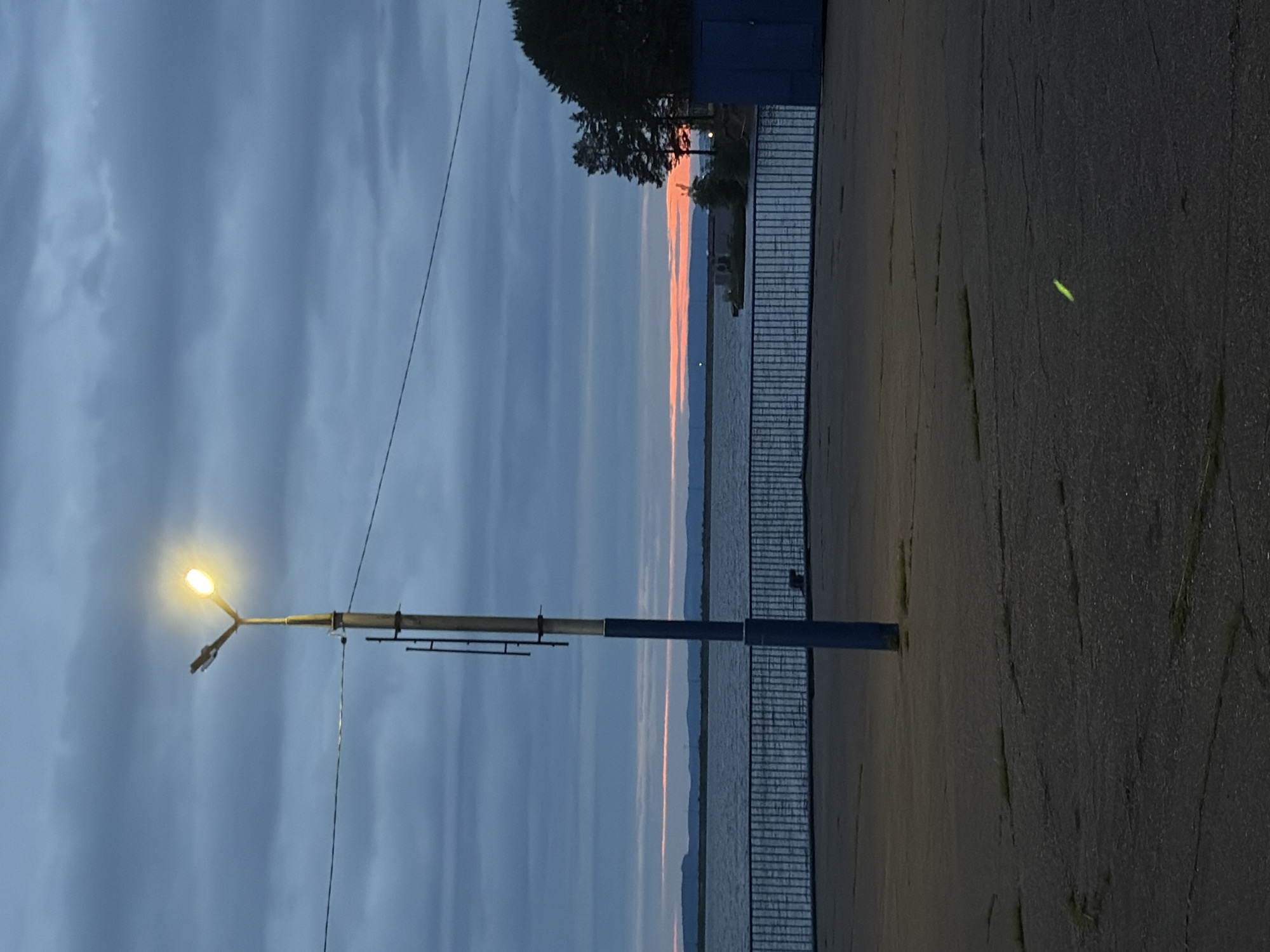 The scene depicts an outdoor waterfront area during twilight or dawn in an unknown location. A tall, illuminated street lamp stands centrally in the foreground, featuring a metallic gray upper section with a ladder-like attachment and a dark blue lower section. The lamp emits a bright yellow light from a fixture with two upward-angled arms. A thin, dark cable or wire spans horizontally across the upper mid-ground, passing behind the lamp post.
The immediate foreground consists of dark gray asphalt pavement marked by numerous cracks and sporadic patches of green vegetation. A faint, glowing green linear object or reflection is present on the pavement in the lower right quarter of the image.
Beyond the pavement, a dark blue metal barrier fence with vertical bars extends horizontally, separating the paved area from a large body of water. The water is dark blue, exhibiting subtle surface ripples, and stretches towards the horizon.
On the distant horizon, a narrow, horizontal band of intense orange and red light is visible, indicative of either sunrise or sunset. Behind this light band, dark, undulating landforms, consistent with distant hills or mountains, are silhouetted against the sky.
To the far right, a cluster of dark green coniferous trees stands on a land extension or shoreline. Adjacent to these trees, a dark blue, rectangular, container-like structure is visible, along with smaller, lighter-colored structures situated closer to the water's edge. The sky above is overcast with blue-gray clouds. No individuals or specific actions are depicted in the image.