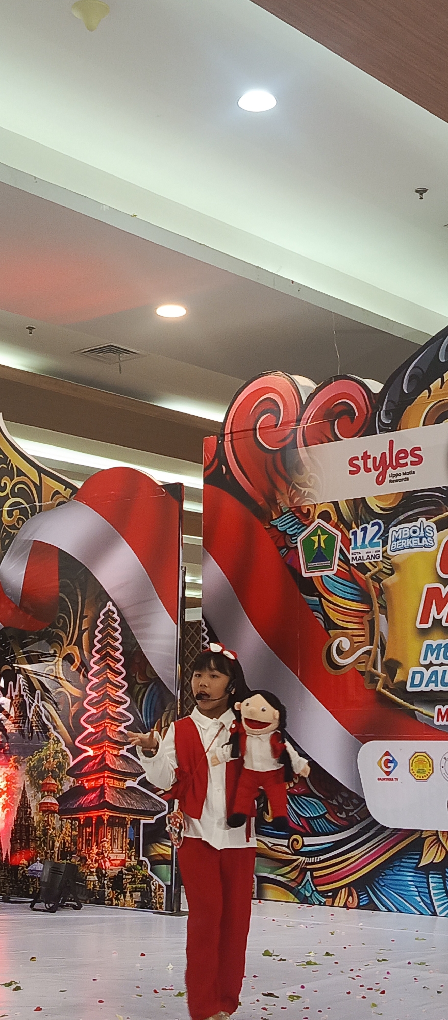 A young girl with a headset is performing on a stage, holding a puppet. She is wearing a white shirt, a red vest, and red pants. The puppet is dressed in a similar outfit. The stage backdrop features a stylized Indonesian flag and imagery of traditional Balinese architecture. To the right, a banner displays "styles" and "Lippo Malls Rewards," along with logos including one for "Malang City." The scene appears to be an indoor event, possibly a mall or cultural center, in Malang, Indonesia. The floor is decorated with scattered red and green confetti.