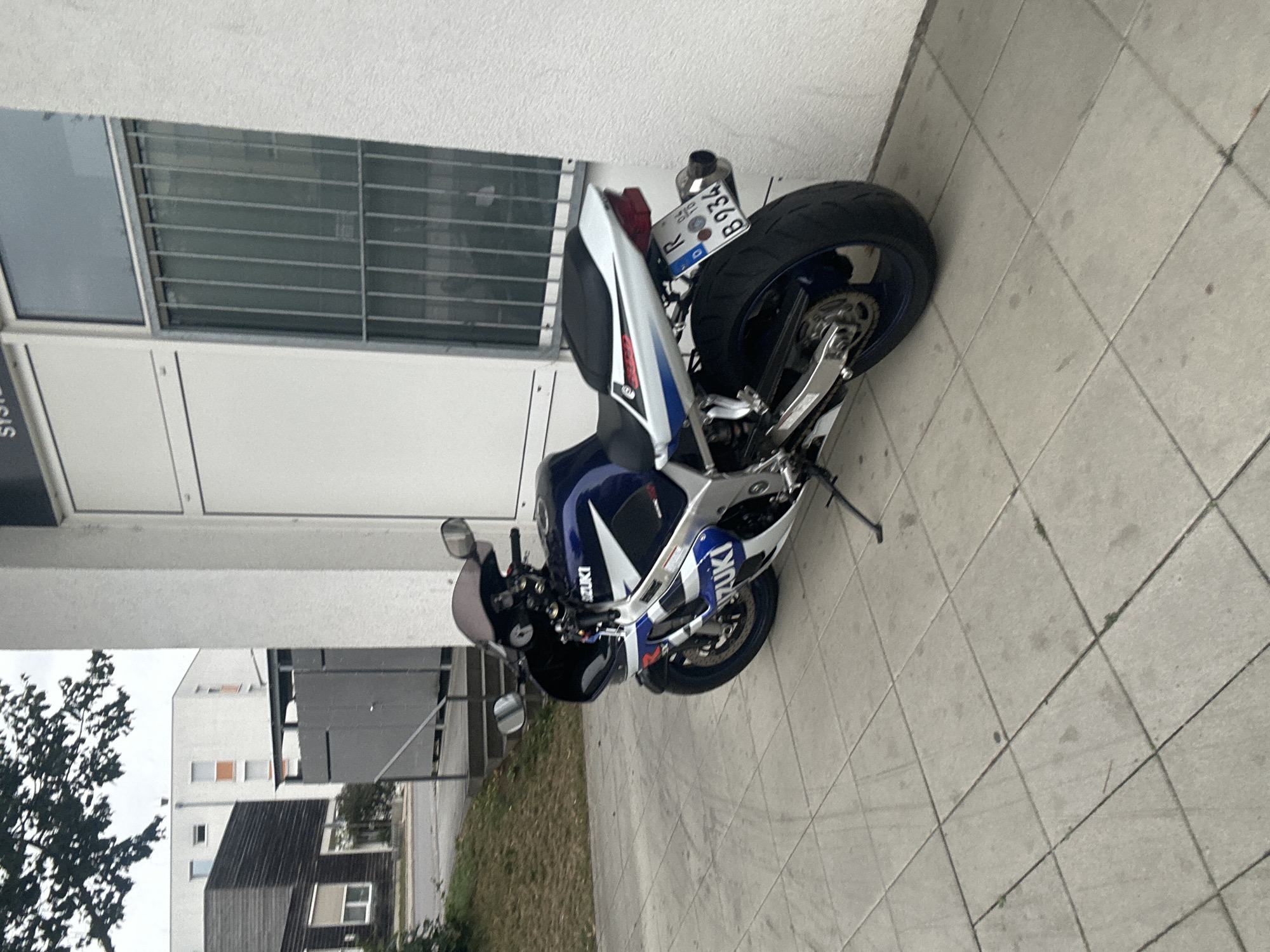 A blue and white Suzuki sport motorcycle is parked on a tiled sidewalk in front of a white building with a barred window. The motorcycle has a license plate reading "R B934". In the background, there are several modern buildings, some with dark siding and others with white facades and orange window frames. Trees with dark green foliage are visible to the left of the scene. The overall environment appears to be an urban setting in Regensburg, Germany.