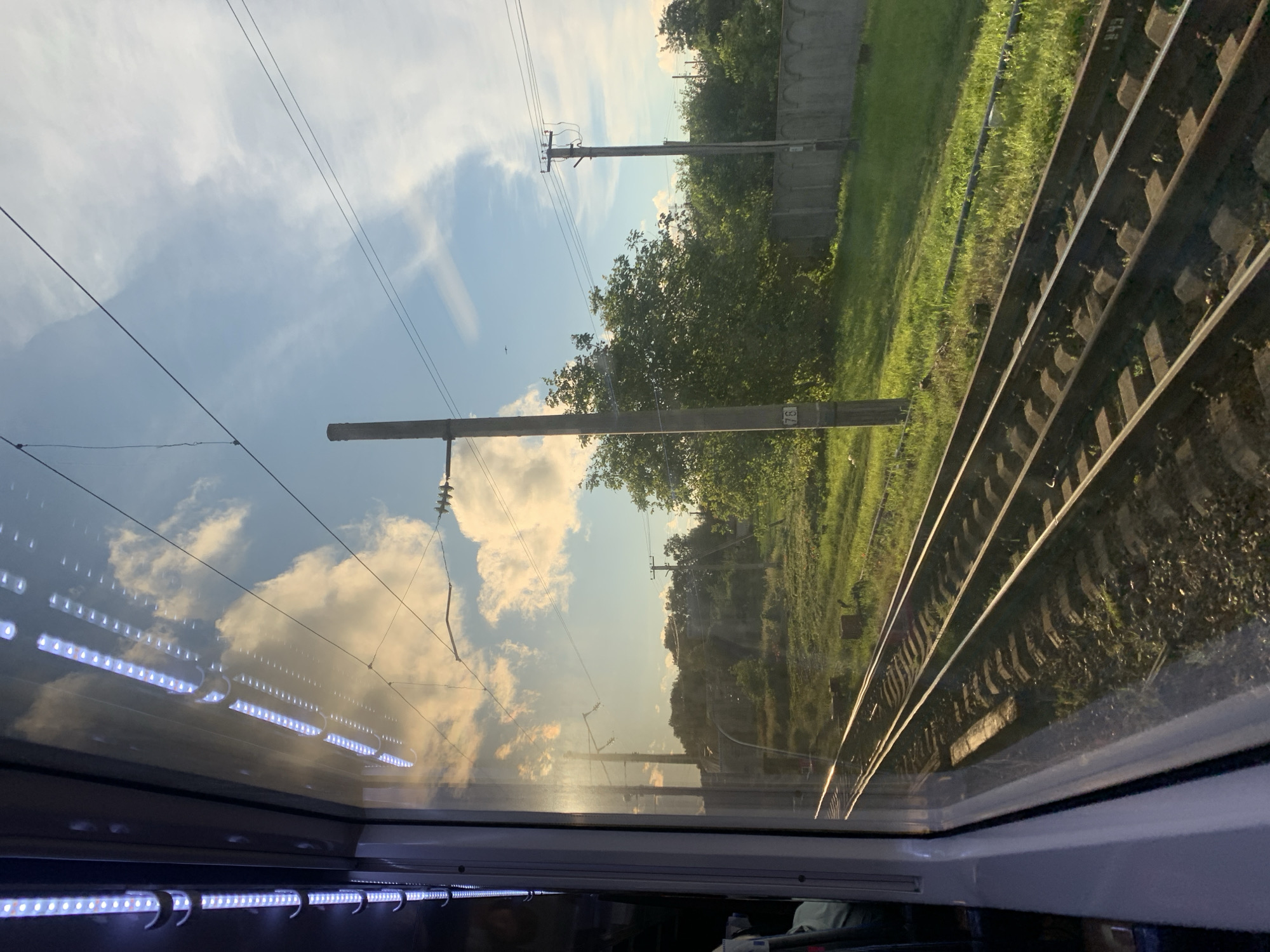 The image depicts a view from inside a train, looking out a window at a rural landscape with railway tracks in the foreground. The tracks run parallel to the train's direction of travel and are flanked by green grass and gravel. In the background, there are trees, power lines, and a concrete fence. The sky is blue with scattered clouds, and the sun appears to be setting, casting a warm glow on the scene. The reflection in the train window shows interior lights and parts of the train's interior. No people are clearly visible. This scene is likely from the vicinity of Marianivka, Ukraine, given the presence of train tracks and rural elements common to the region.