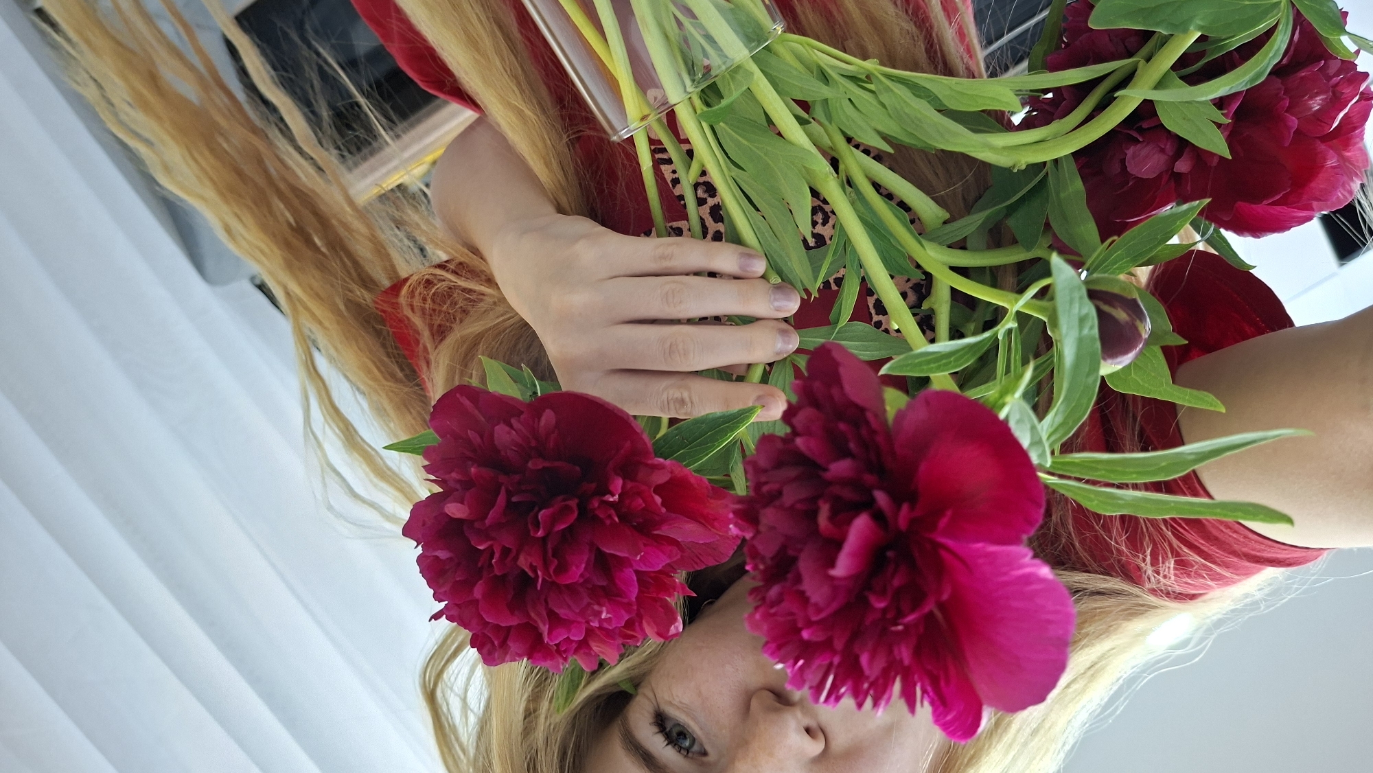 The image depicts a young woman in an overhead shot, with her body oriented horizontally across the frame. Her face, showing one light-colored eye, part of her nose, and mouth, is visible on the right side. She has long, wavy blonde hair that flows upwards towards the top left of the frame. Her left hand, with fingernails painted in a light neutral shade, gently holds a clear glass vase filled with dark pink/magenta peonies.
The bouquet contains several fully bloomed peonies and numerous green stems with leaves. Two large, vibrant peonies are prominently featured in the foreground, positioned near her face. The woman is wearing a red top, and a subtle leopard print pattern is visible beneath the red fabric, possibly as an underlayer or part of the garment's design.
The activity appears to be a staged portrait, with the woman holding and possibly admiring the flowers. The background on the left consists of a light-colored surface with distinct vertical lines, resembling window blinds or wall panels. A darker, indistinct area is visible in the upper right background. The lighting is soft and natural, suggesting an indoor setting during daylight. The visible part of her face conveys a calm and gentle expression. There is no visible text. The location context for this scene is Chaikovsky, Russia.