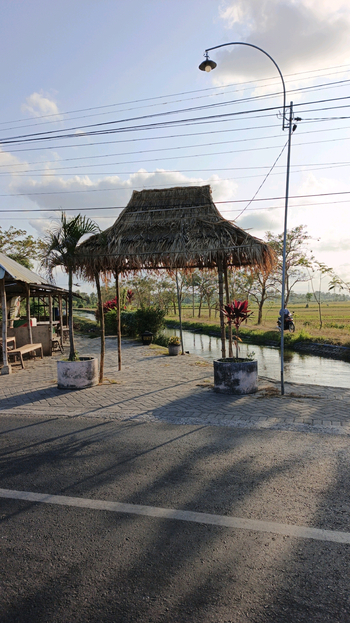 The image shows a street scene in Sanggrahan, Indonesia, with a thatched-roof pavilion situated on a paved area next to a road. A narrow canal or waterway runs parallel to the paved area, bordered by a rice field. In the distance, there are trees and a clear sky with scattered clouds. A motorcycle is visible in the background on the right side of the image. A lamppost stands near the pavilion, and power lines are strung overhead. The foreground is dominated by the asphalt road, with shadows of the pavilion and surrounding elements cast upon it. The overall impression is of a rural or semi-rural setting, possibly a rest stop or market area, at dusk or dawn given the lighting.