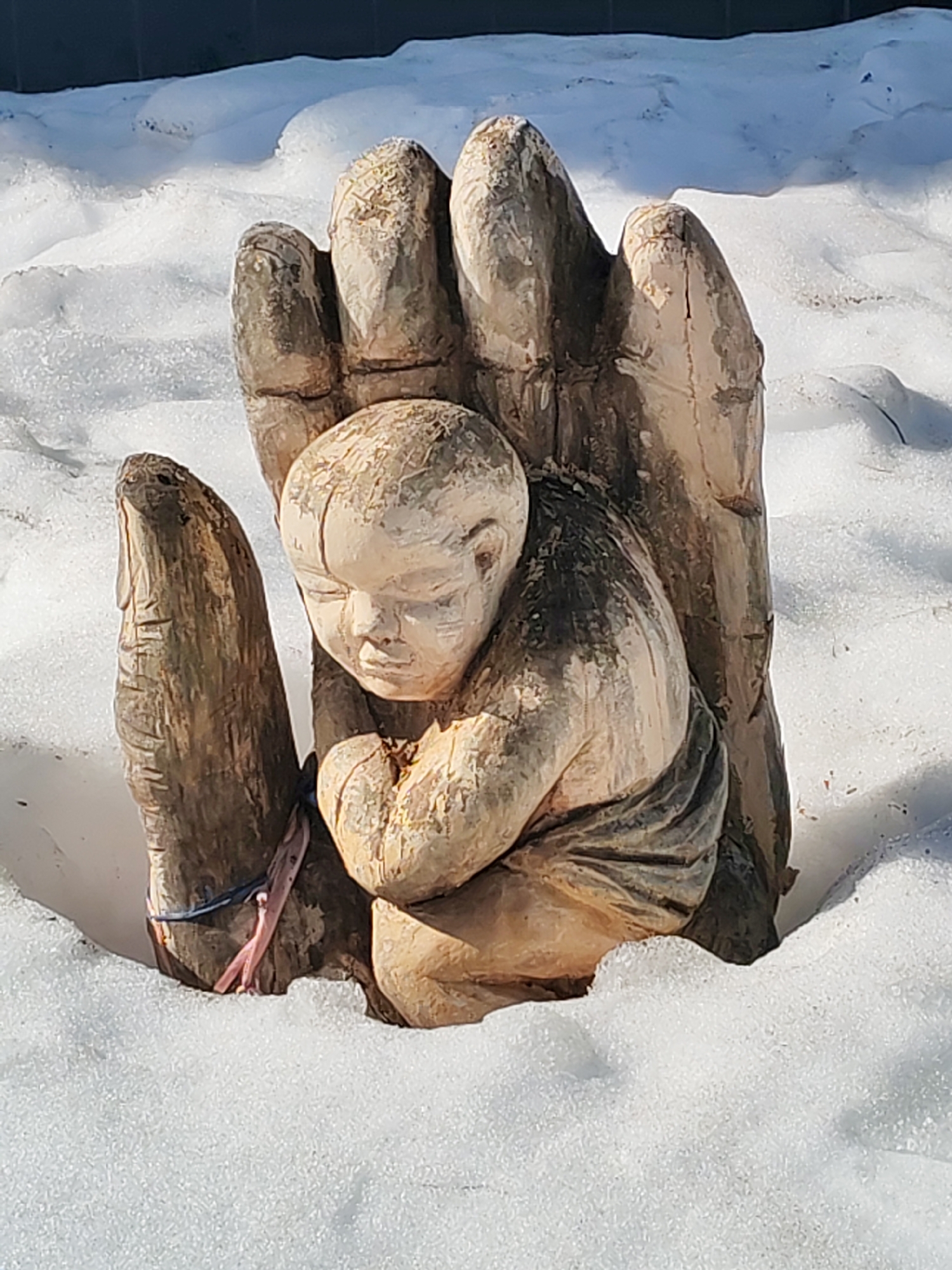 The image shows a wooden sculpture of a hand cradling a child, set in a snow-covered area. The sculpture is located outdoors, likely in a public space. It is daytime, with strong sunlight casting shadows on the snow and sculpture. The hand is large and stylized, made of rough-hewn wood, with clearly defined fingers. The child is depicted in a fetal position, nestled within the palm of the hand. A small blue and pink ribbon adorns one of the fingers of the hand. The sculpture is embedded in a blanket of snow, suggesting it has been in place for some time during winter conditions. The location is Chaikovsky, Russia.