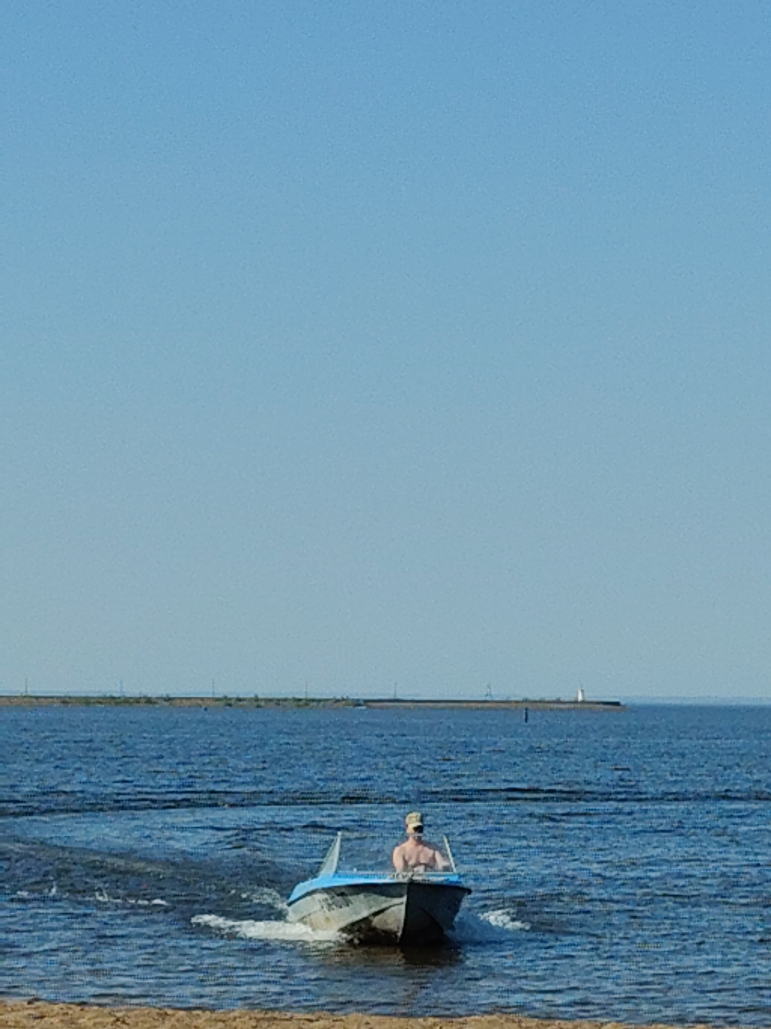The image depicts a person in a motorboat on a body of water, possibly a lake or a bay. The person appears to be operating the boat. In the background, there is a landmass with a structure that resembles a lighthouse. The sky is clear and blue. The location is unknown.