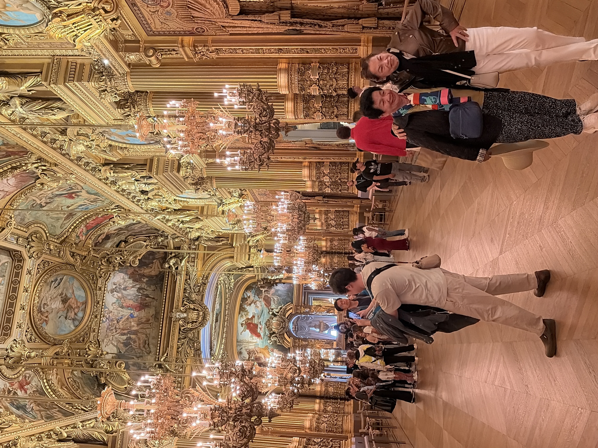 The image captures an interior view of the Palais Garnier opera house in Paris, France. The opulent Baroque architecture is evident with its ornate golden decorations, detailed moldings, and grand ceiling frescoes. Large crystal chandeliers hang from the ceiling, illuminating the vast hall. Several people are present, appearing to be tourists or visitors, some taking photos. In the foreground, two women are posing for a picture, smiling. Other individuals are seen walking or standing in the background, engaged in various activities. The floor is made of polished wooden planks arranged in a parquet pattern. The overall ambiance suggests a historical and luxurious setting.