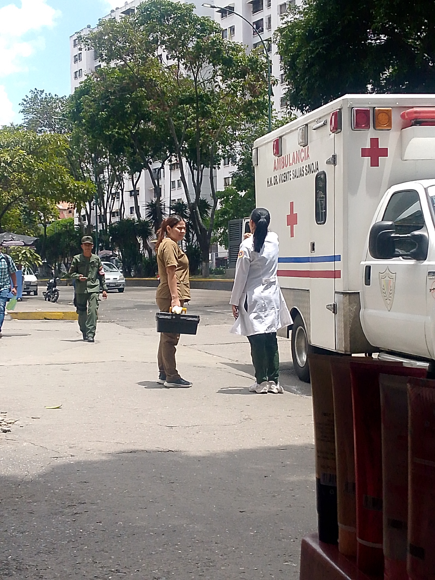 The image shows a street scene in Caracas, Venezuela. An ambulance is parked on the right side of the frame. "Ambulancia" and "H.M. Dr. Vicente Salias Sanoja" are written on the side of the ambulance. A woman in a white lab coat is standing beside the vehicle. Another woman in a brown uniform is standing on the street, holding a black container. A soldier in green uniform is walking towards them. Other people and vehicles are visible in the background. The environment includes trees and a building in the background. The foreground contains part of a shelf with what appears to be product packaging.