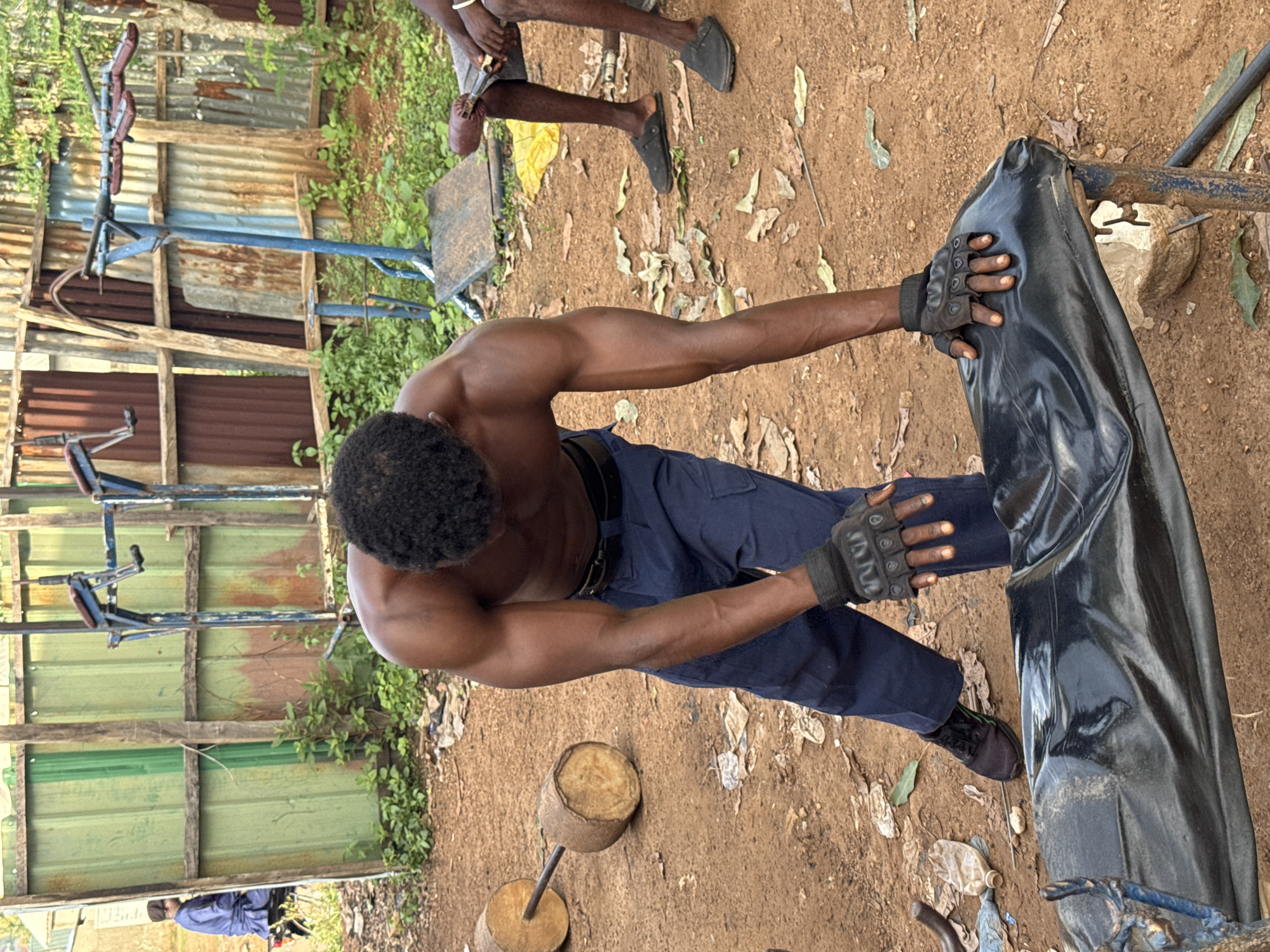 In Akwanga, Nigeria, a shirtless Black man with dark skin and short, curly hair is performing a workout in an outdoor gym. He is wearing dark blue pants, black gloves, and black athletic shoes. He is bending over a black, tarp-covered bench, with his hands resting on the tarp. In the background, there is a makeshift weightlifting apparatus with blue metal supports and brown padding, as well as a barbell made of logs and a metal rod. The ground is dirt and scattered with debris, including plastic bottles and leaves. To the left of the image, another man in a blue robe and head covering is sitting on a motorcycle, facing away from the camera, with a green corrugated metal fence behind him. The overall environment appears to be a modest, rustic outdoor fitness area.