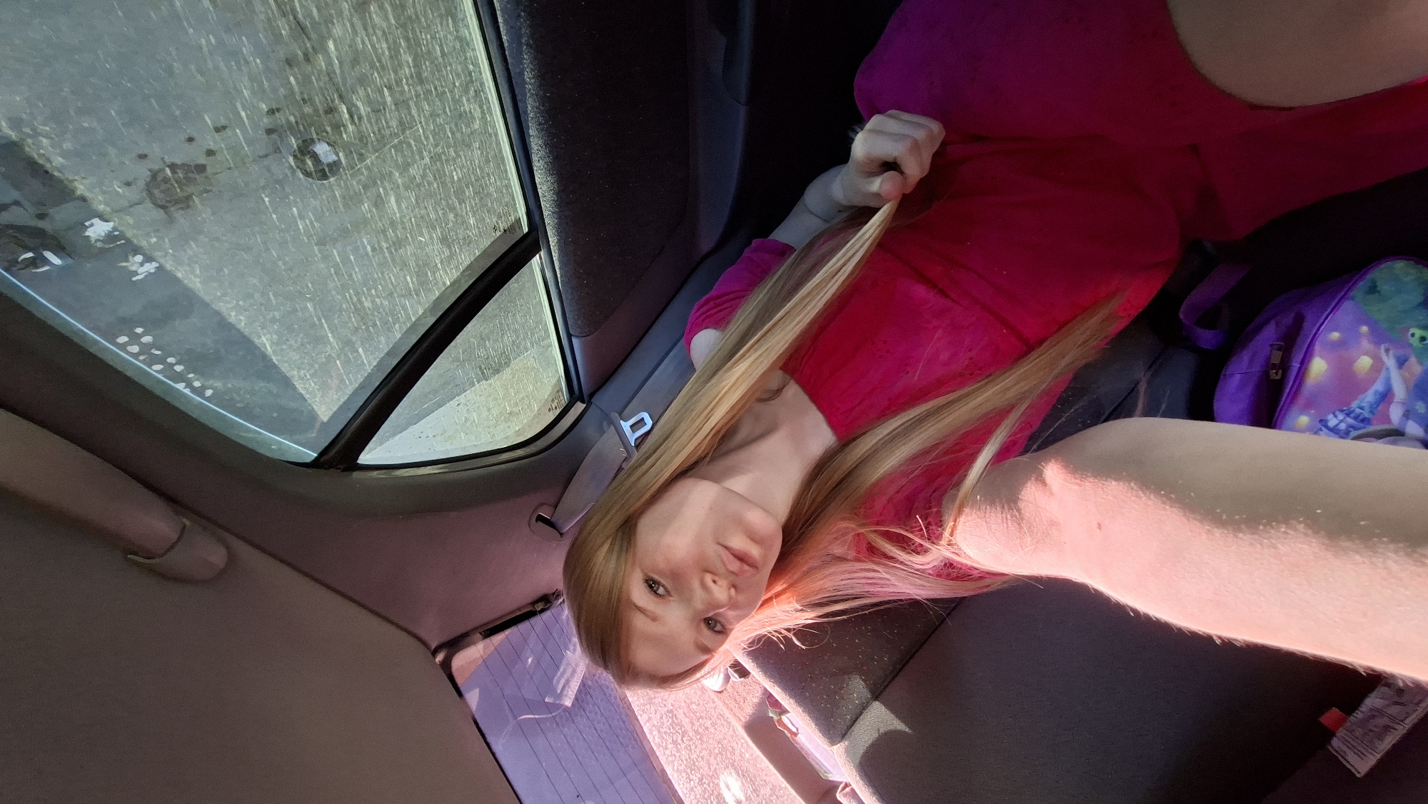 A young woman with long blonde hair, wearing a bright pink off-the-shoulder dress, sits in the back seat of a car. She is looking at the camera, puckering her lips. A purple backpack with a cartoon character design is visible next to her. The car's interior, including the grey upholstery of the seat and the tinted window, is visible. The exterior view through the window appears to be a dusty, outdoor environment. The image was taken on a sunny day.