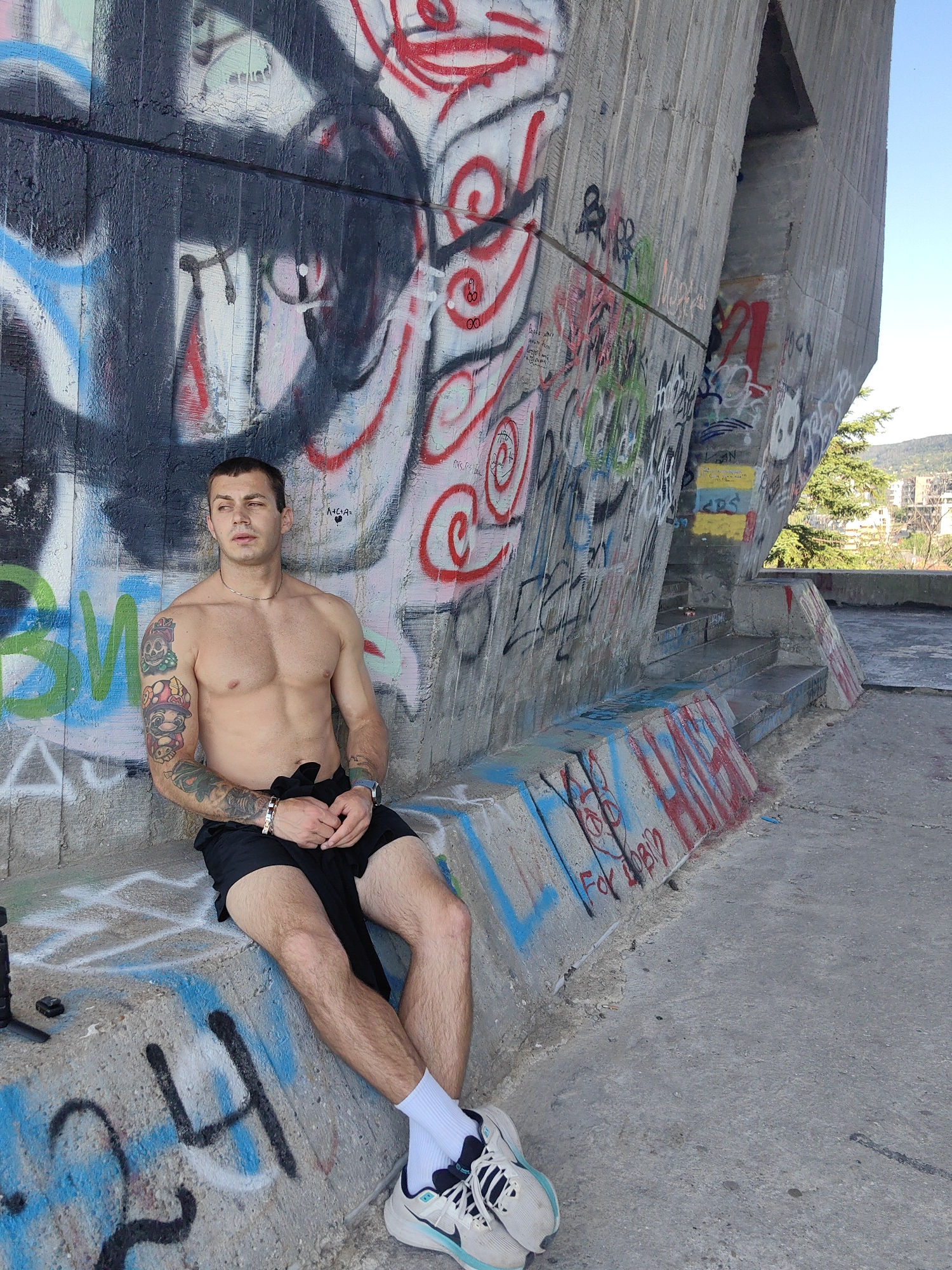 A shirtless man with tattoos on his left arm sits on a concrete ledge in front of a graffiti-covered wall. He is wearing black shorts, white socks, and athletic shoes. His hands are clasped in his lap, and he is looking to his right. The wall behind him is covered in various graffiti tags and drawings, with a bright blue sky visible above. The scene appears to be an outdoor urban environment, possibly an underpass or a construction site, given the concrete structures and the presence of graffiti.