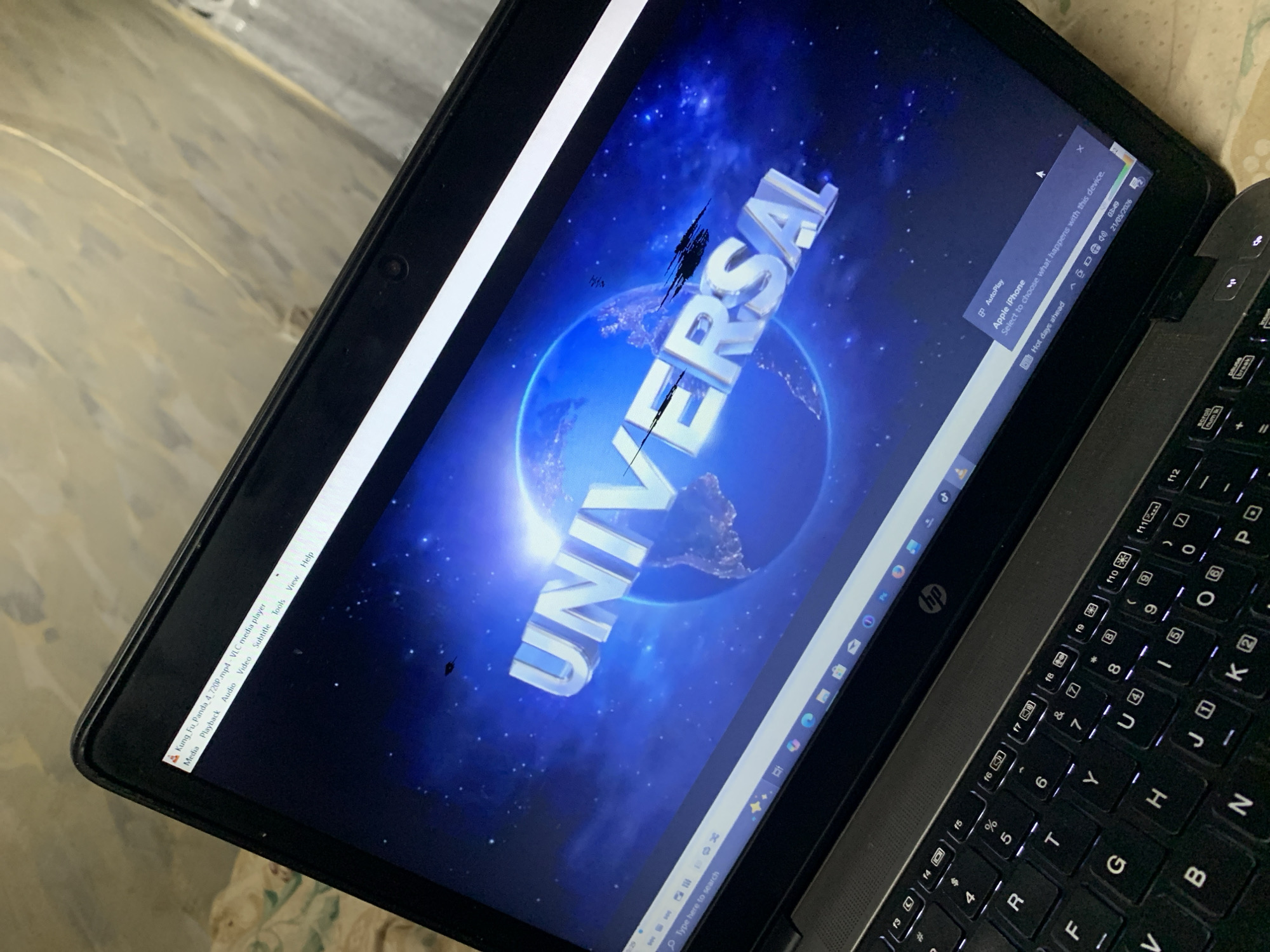 The image shows a laptop screen displaying the Universal Studios logo. The keyboard of the laptop is visible, along with a pop-up window suggesting connection to an Apple iPhone. The scene is not located in Rafin Par, Nigeria.
