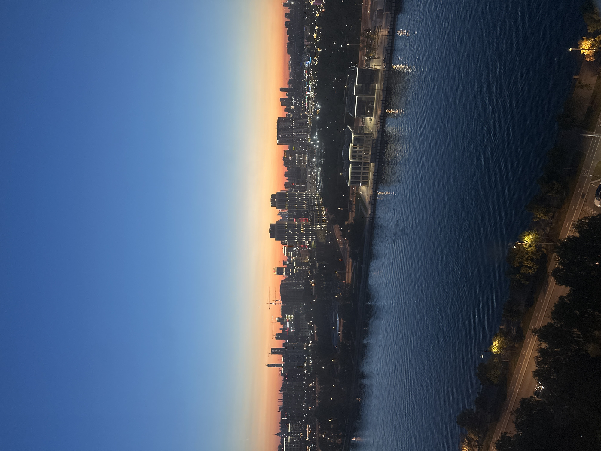 The image depicts a cityscape at twilight, with a wide river in the foreground and a sprawling urban landscape beyond. The sky transitions from a deep blue at the top to a vibrant orange and pink near the horizon, indicating the last moments of sunset or the first moments of sunrise.
The river occupies the lower half of the image and is a dark, deep blue, reflecting the lights of the city and the sky. Small ripples are visible on its surface, suggesting a gentle current. Along the riverbank, there is a paved promenade with modern, minimalist buildings featuring illuminated windows and facades. Further inland, a dense band of trees separates the river from the city skyline.
The city skyline is dominated by a variety of buildings, including skyscrapers and lower-rise structures. Many buildings are illuminated, casting a warm glow and creating a sense of a bustling urban environment. Construction cranes are visible among some of the taller buildings, suggesting ongoing development. Streetlights and lights from vehicles are also visible, lining roads and pathways within the city.
The foreground of the image shows a tree-lined road running parallel to the river. Streetlights illuminate the road and the surrounding trees, casting long shadows. A few cars are visible on the road, one of which has its headlights on. The trees are dark silhouettes against the illuminated road and the distant city lights.
Based on the architectural style, the presence of construction cranes, and the overall urban layout with a prominent river, this scene is likely located in Moscow, Russia. The buildings and the river's expanse are consistent with areas along the Moskva River, such as the Moscow City business district or the areas around the Luzhniki Stadium. The modern architecture of the buildings along the riverfront suggests a recently developed or redeveloped area.