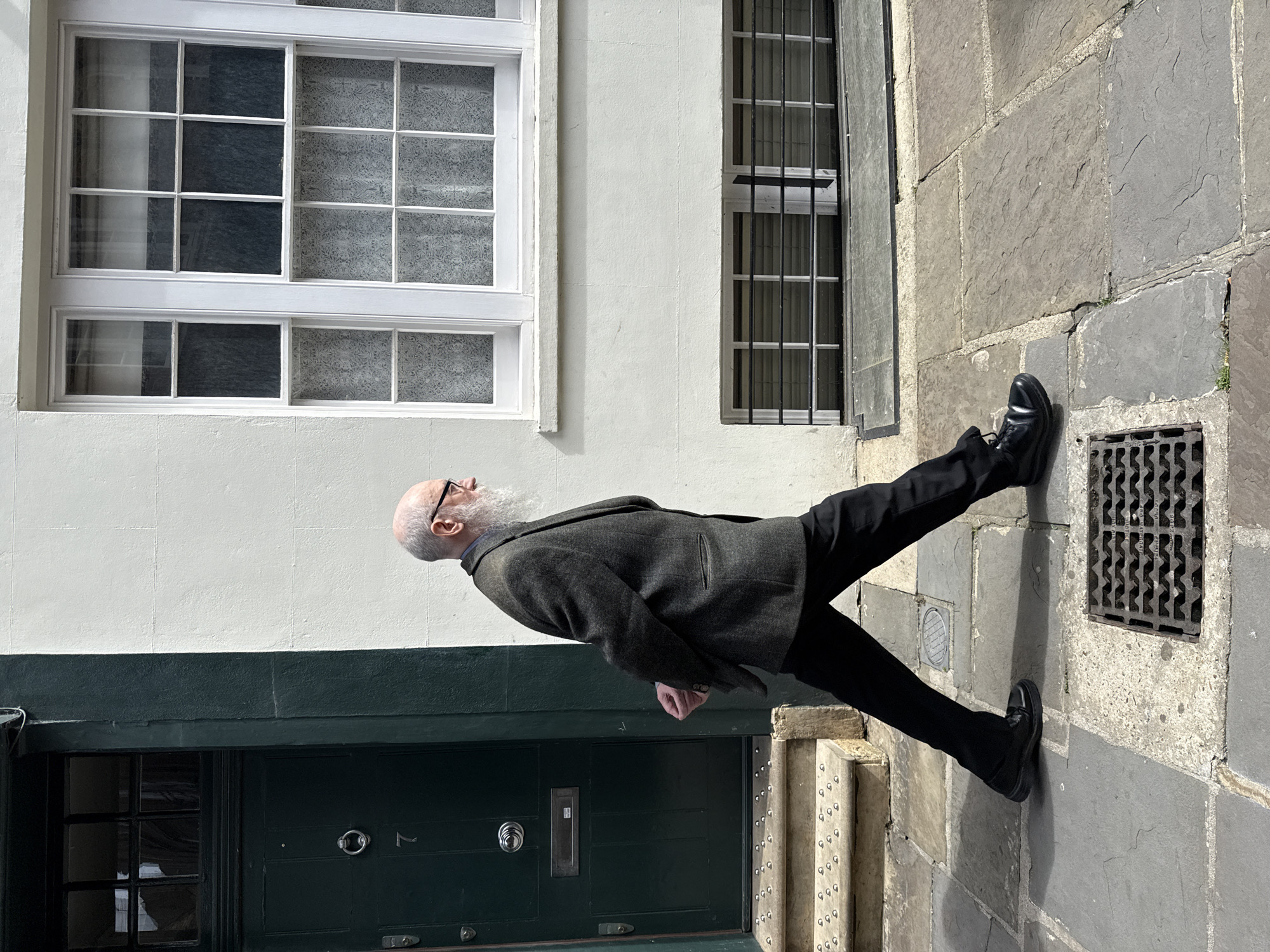 This outdoor, eye-level, full-length photograph, set in London, United Kingdom, captures an elderly man standing on a stone pavement in front of a building facade.
The man, bald with a long, full white beard and glasses, is dressed in a dark grey, textured jacket, a dark shirt with a visible collar, black trousers, and black dress shoes. He is positioned facing left in the frame, with his head tilted upwards, suggesting he is looking at something high on the building or in the sky. His hands are clasped or held together in front of his waist.
The building behind him features a white upper section and a contrasting dark green lower section. In the white part of the facade, two large white-framed windows with multiple panes are visible. Some panes reflect the surroundings, while others are covered with a subtle, patterned privacy film. To the right, a narrower, vertical window with dark metal security bars adds another architectural detail.
At the ground level, a dark green front door is prominent, featuring a silver round doorknob and a silver rectangular letterbox plate. The house number "7" is clearly visible next to the doorknob. The pavement consists of large, irregular grey stone slabs. A rectangular metal drain cover with a grid pattern and some indistinguishable text is embedded in the pavement near the man's right foot, with another smaller, round metal cover further back.
The scene is brightly illuminated by natural daylight, indicating a sunny day, likely midday or early afternoon, given the clear, sharp shadows cast. The man appears to be in a moment of quiet observation or contemplation in this urban setting.