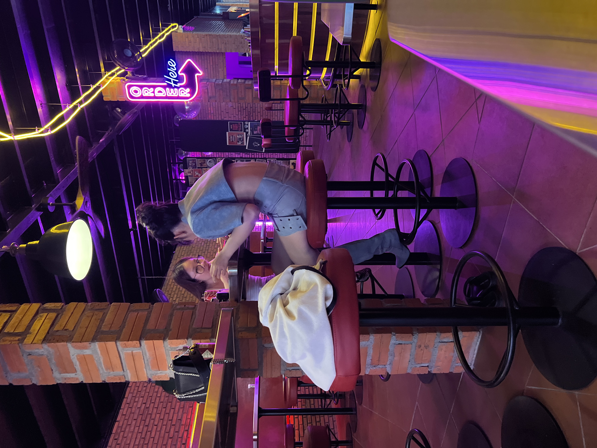 Inside a brightly lit bar in Bac Nha Trang Ward, Vietnam, two people are seated at a high bar. One person, wearing a blue outfit and gray boots, sits on a red bar stool. The other person is positioned closer to the bar. Overhead, a neon sign with the word "Order" and an arrow pointing up is visible. The bar area is illuminated with both yellow and purple neon lights, creating a vivid atmosphere. A black purse hangs near the bar. The bar stools are lined up along the bar counter. The walls have a brick pattern.