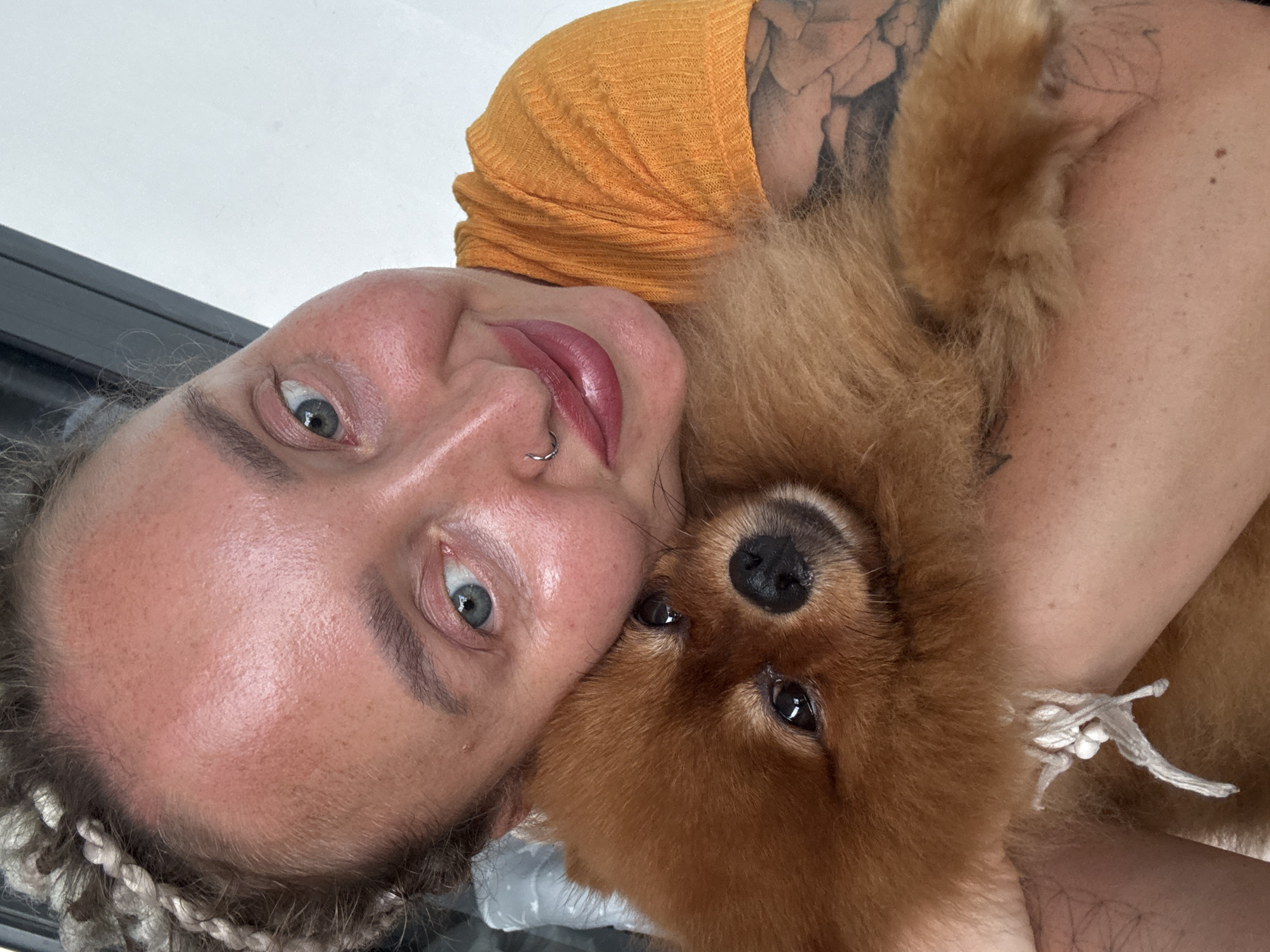 In this close-up selfie, a woman with blonde braided hair is embracing a small, fluffy brown dog. The woman is wearing an orange t-shirt and has a nose ring. Her skin appears flushed, and she has makeup on her lips. The dog is nestled against her face, with its eyes looking towards the camera. The background is a plain white wall. The setting is indoors, likely a home or a private residence, with no visible indications of being in a specific city like Bac Nha Trang Ward, Vietnam.