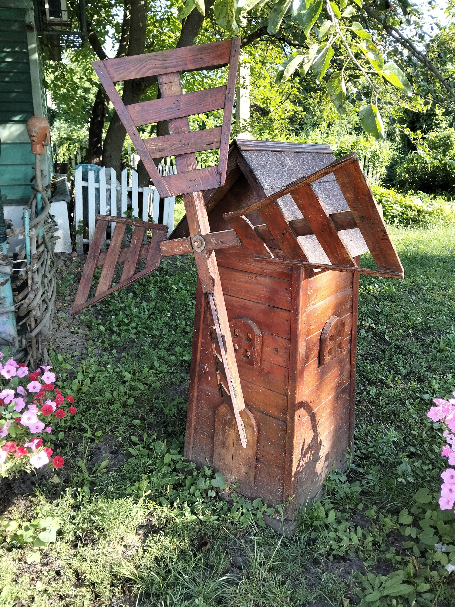 This image, taken in Kliushnykivka, Ukraine, depicts a rustic, wooden windmill decoration situated in a garden. The windmill is constructed from stained wood, with a weathered appearance. It features a central body with two small, arched windows and a pitched roof covered in gray shingles. Two large, fan-like blades, also made of wood, are attached to a central shaft that extends from the body of the windmill. One blade is angled upwards, while the other is angled downwards and slightly to the left. A wooden arm extends from the shaft downwards, near the base of the windmill.
The windmill stands on a patch of uneven ground covered in green grass and various low-lying plants, with some exposed soil visible. To the left of the windmill, a cluster of pink and red petunias is in bloom. Behind the windmill, a white picket fence is visible, and beyond that, a dense green foliage of trees and bushes. To the far left, a portion of a teal-colored building with horizontal siding and a decorative terracotta pot on a rustic wooden post is partially visible. The lighting suggests it is daytime, with sunlight casting shadows on the windmill and the surrounding ground.