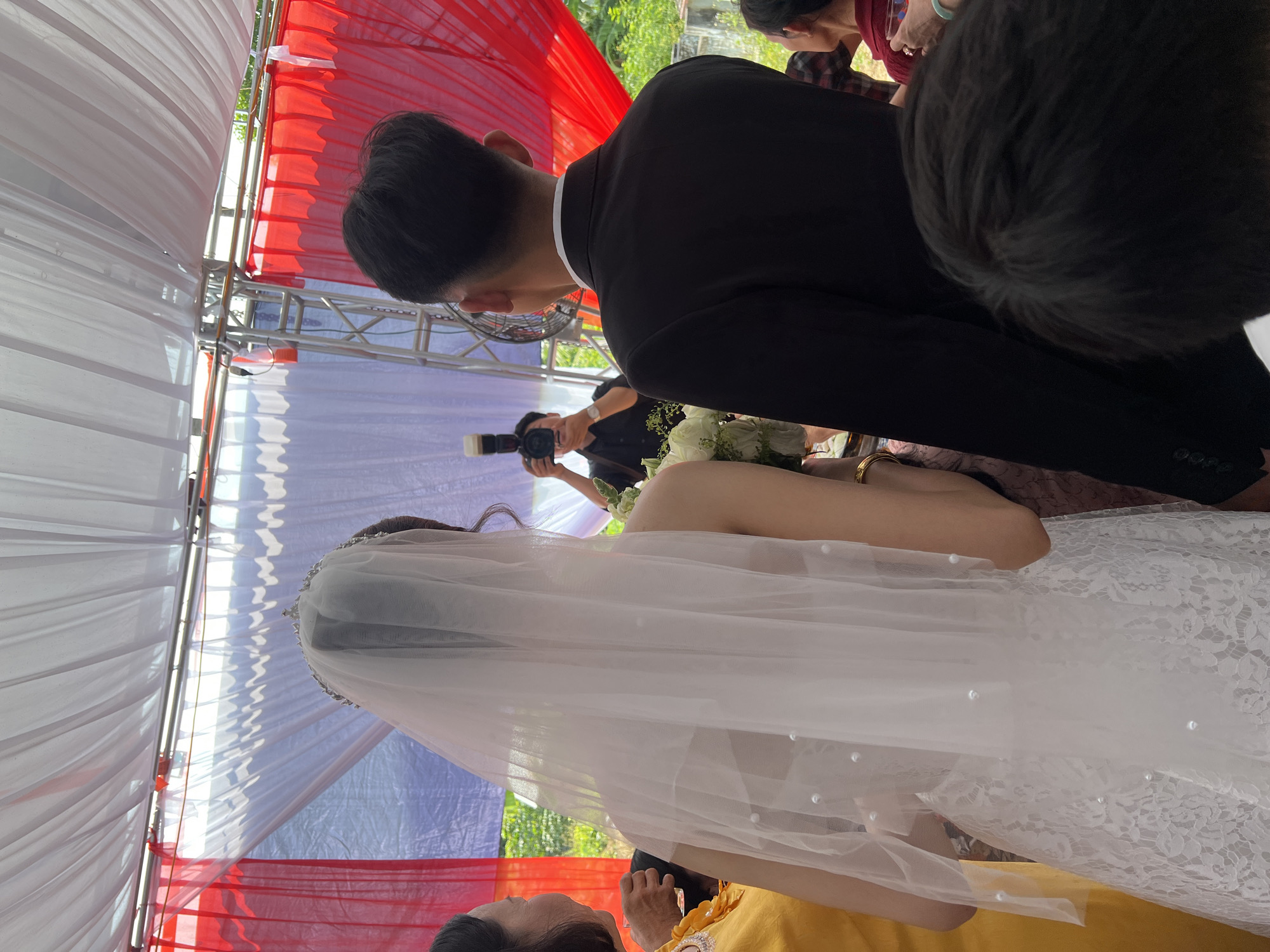 This image captures a moment at a wedding ceremony or reception in Tay Son Commune, Vietnam, observed from behind the bride and groom. The setting is under a decorated tent or canopy.
The main subjects are the groom, seen from the back and left side, wearing a black suit jacket over a white collared shirt, and the bride, positioned to his right, also viewed from behind. The bride is adorned in a white, off-the-shoulder wedding dress featuring lace detailing. A long, sheer white veil, embellished with small pearl-like beads, cascades over her back. She holds a white floral bouquet, partially visible between her arm and the groom.
In the background, a person, likely a photographer, is visible holding a black camera with an attached flash unit, actively capturing the event. Other attendees are partially obscured around the edges of the frame.
The event takes place within a structure made of metal trussing, draped with panels of white and vibrant red fabric, indicating a festive occasion. Bright natural light filters through the canopy, suggesting it is daytime. Green foliage is visible in the distance beyond the canopy, indicating an outdoor or semi-outdoor location. No visible text is present in the image.