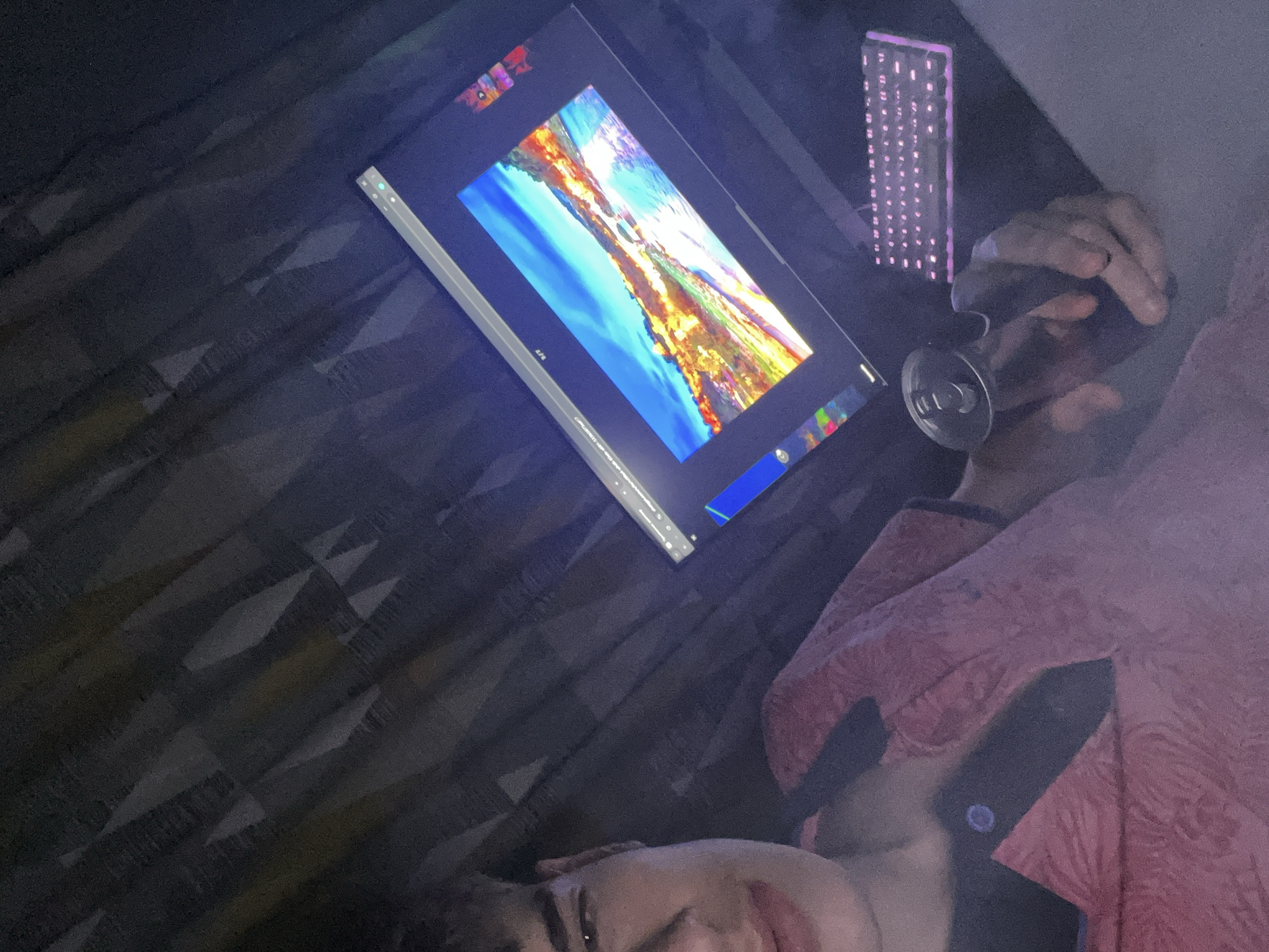 A photograph taken indoors shows a person in partial view, lying down and angled towards a monitor screen displaying an image reminiscent of a landscape painting with vibrant colors. The monitor is positioned on a surface with a dark, patterned texture. The person is holding a metallic can, possibly a beverage, and a gaming keyboard with illuminated keys is visible in the background. The image suggests a casual, relaxed setting, possibly the person's private space. There is no location in the city of Unknown, Unknown, as none can be determined from the image.
