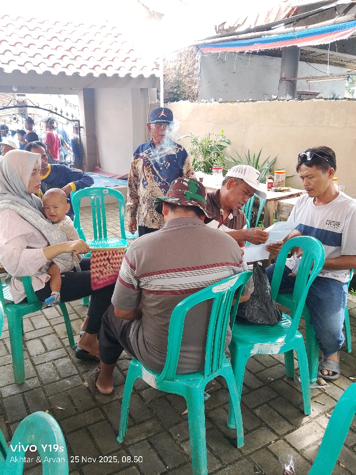A group of people are seated in a semi-outdoor setting. A woman wearing a hijab is holding a baby. An older man in a patterned shirt and baseball cap is smoking. Three men are seated at a table, looking at papers. All are seated on matching teal plastic chairs. A building is in the background. A banner with red, white, and blue stripes is hanging on the building.