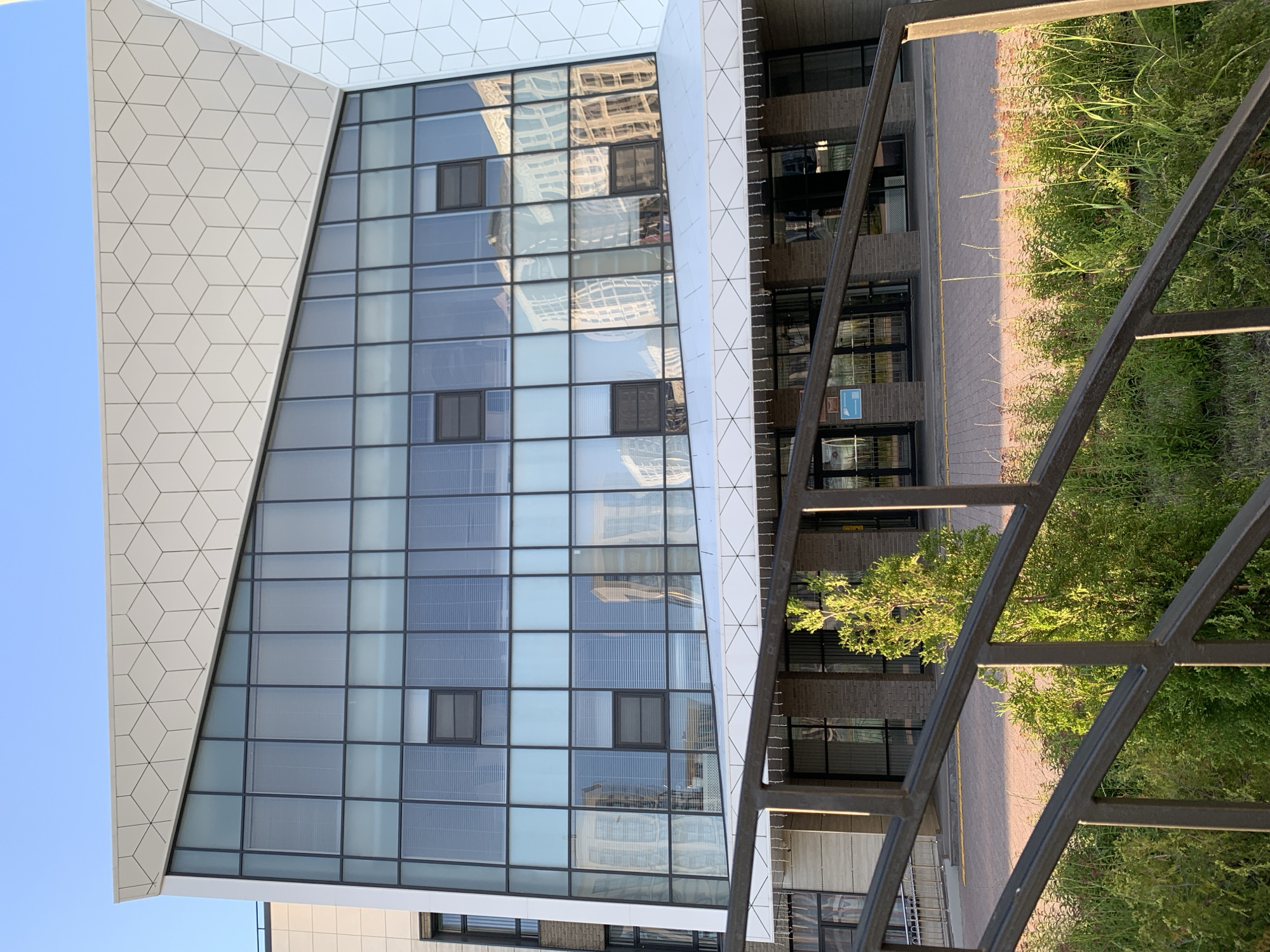 The image shows the exterior of a modern building with a distinctive angled facade. The upper portion of the building features a large expanse of glass windows, reflecting surrounding structures. Above the windows, a white paneling with a geometric cube pattern is visible. The lower level of the building has a brick facade and glass doors. In the foreground, a dark metal railing with a crisscross design is in focus, with green vegetation and a small tree below it. The scene appears to be outdoors, under a clear blue sky. The location is identified as Stavropol, Russia.