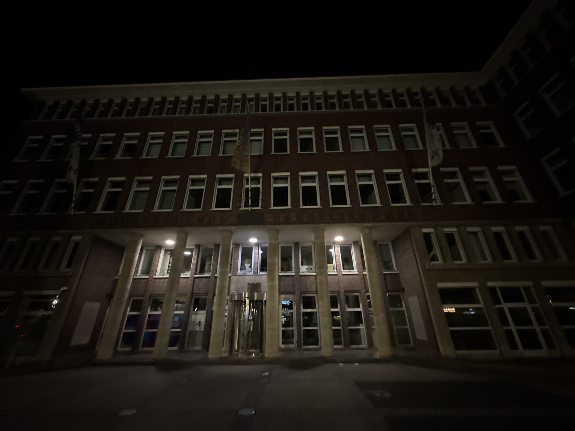 A wide-angle, low-light photograph captures the exterior of a multi-story brick building at night in Duisburg, Germany. The building features a rhythmic pattern of windows on multiple floors, with the upper floors having smaller, recessed windows and the lower floors displaying larger, more prominent window panes. A prominent revolving glass door is visible at street level, flanked by large columns. A Ukrainian flag is displayed on a flagpole in front of the building. The street in front of the building appears to be paved. The overall scene suggests an official or institutional building, illuminated by artificial light sources within and around the structure.