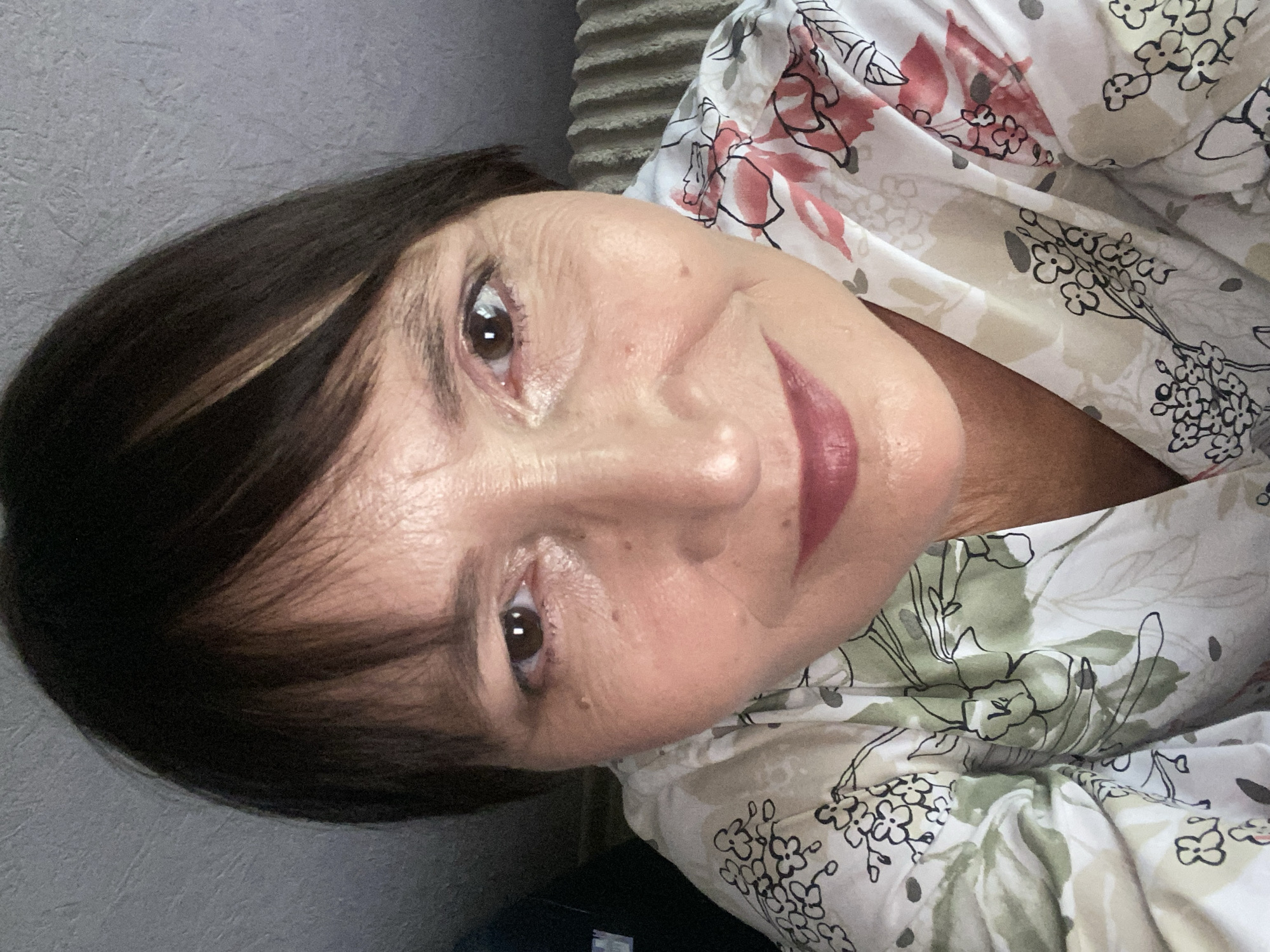 A close-up, low-angle, selfie-style photograph shows the upper torso and face of a woman. She has dark brown hair with some lighter streaks, and her bangs are swept to the side. Her eyes are brown, and she is smiling faintly. She is wearing a white shirt with a floral pattern of green leaves and pink flowers. The background is blurred and appears to be an indoor setting with a textured wall and the ribbed back of a couch. There is no discernible information to place the image in Novokuznetsk, Russia.