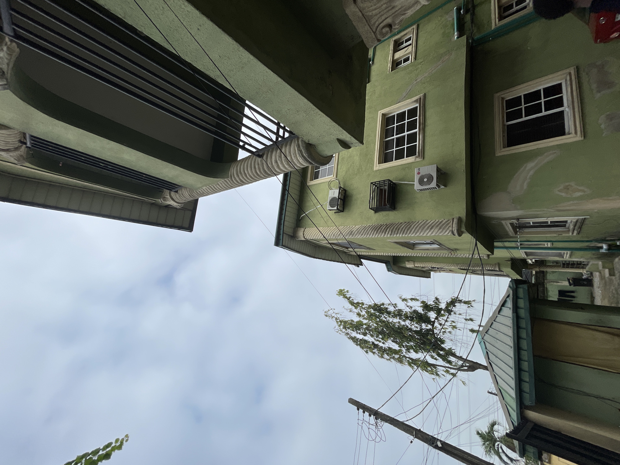 The image depicts a street scene in Ado, Nigeria, viewed from an upward, tilted perspective. The architecture is dominated by buildings painted in shades of green, featuring rectangular windows and air conditioning units affixed to their facades. Utility poles and a web of electrical wires crisscross the sky. A tree with sparse foliage leans into the scene. The sky is overcast with white clouds.