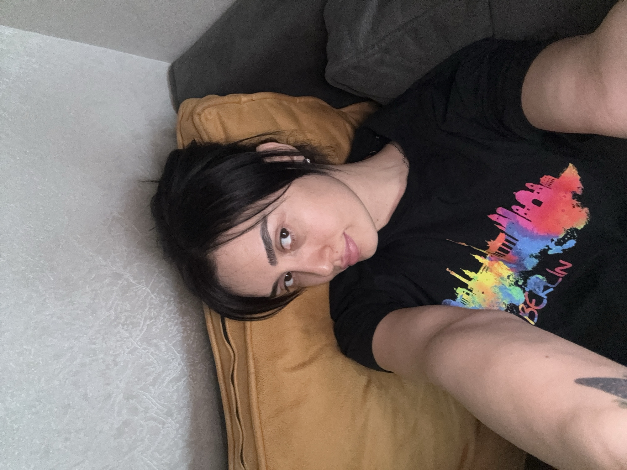 A person lies on a mustard-colored cushion that appears to be part of a sofa, with a gray sofa cushion visible above the mustard one. The person has short, dark hair and is wearing a black t-shirt with a colorful depiction of the Berlin skyline. A tattoo is visible on the person's arm. The background consists of a textured, light gray wall. There is no indication of the location being in Tbilisi, Georgia.