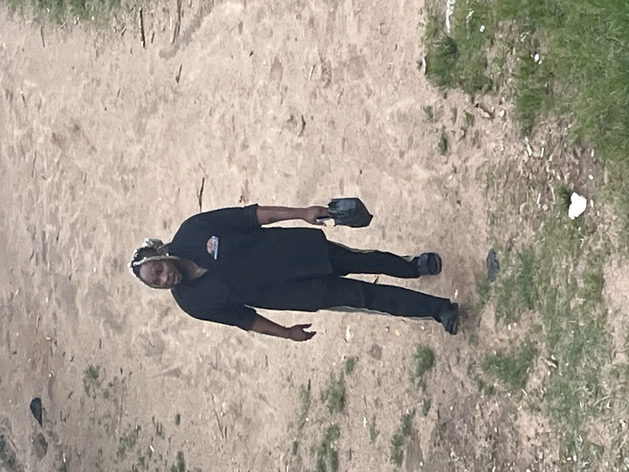 A man with blonde-streaked dreadlocks walks on a dirt path in a grassy area. He is wearing a black short-sleeved polo shirt with a circular logo on the left chest and black pants with a white stripe down the side. He is carrying a small, dark-colored bag in his right hand and wearing dark-colored shoes. The ground is a mix of dirt and sparse green grass, with scattered debris. The background is out of focus, showing more of the dirt path and vegetation. The scene appears to be in an outdoor, unpaved area, possibly a park or a less developed section of the city. There are no other people visible in the image. The lighting suggests it could be daytime. This location could be within the city of Jos, Nigeria, given the description of an outdoor, unpaved, and grassy area.