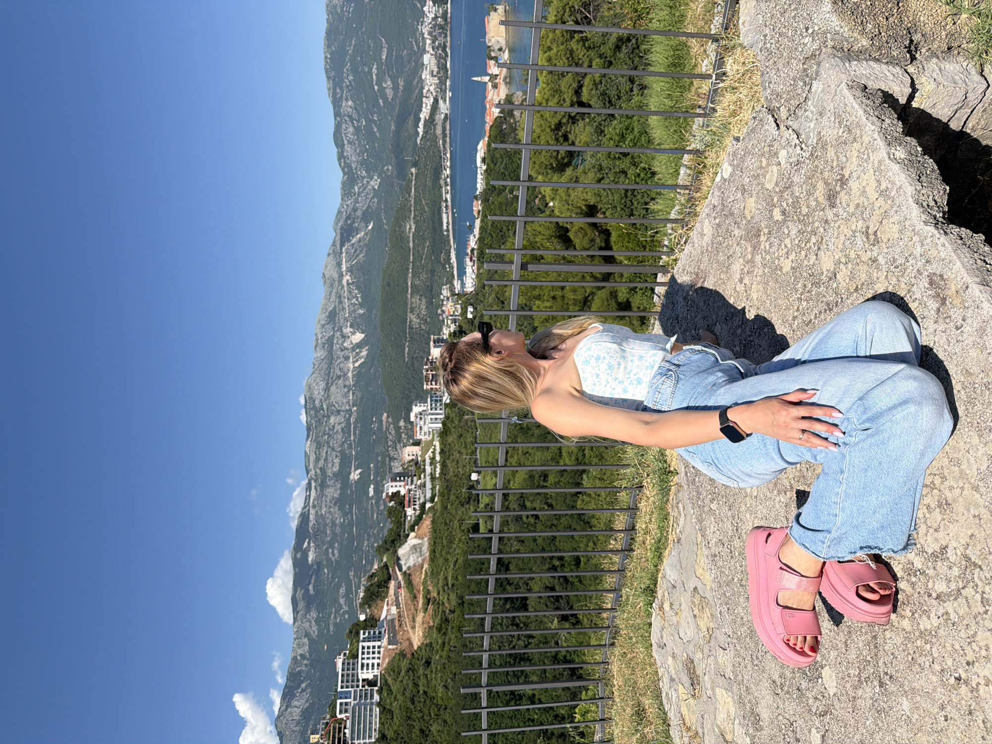 An outdoor, wide-angle shot captures a female kneeling on a stone surface, facing a coastal cityscape and mountain range under a clear blue sky.
The subject is a woman with long blonde hair, wearing a white and light blue floral strapless top, light blue distressed jeans, and pink platform sandals (with "UGG" visible on the strap of the left sandal). She has a dark smartwatch on her left wrist, light pink nail polish on her fingers and toes, and dark sunglasses resting on her head. She is positioned on a flat, light grey, uneven stone slab in the foreground, with her body angled slightly right and her head turned to her right, looking into the distance.
A dark grey metal fence with regularly spaced vertical bars extends horizontally behind the woman, separating the immediate foreground from the view beyond. Directly behind the fence is a dense expanse of green trees and foliage, sloping downwards.
In the middle ground, an azure blue bay is visible, populated by several white boats. A distinct, fortified peninsula, characterized by closely packed buildings and a prominent light-colored church tower, extends into the bay, identifying the location as Budva Old Town, Montenegro. Modern multi-story buildings with white facades are scattered across the green hillsides surrounding the bay.
The background features a dramatic range of rugged, grey-green mountains stretching across the horizon under a brilliant, cloudless blue sky, with a few small white clouds appearing over the left side of the mountain range. The scene is illuminated by bright daylight, suggesting sunny weather.