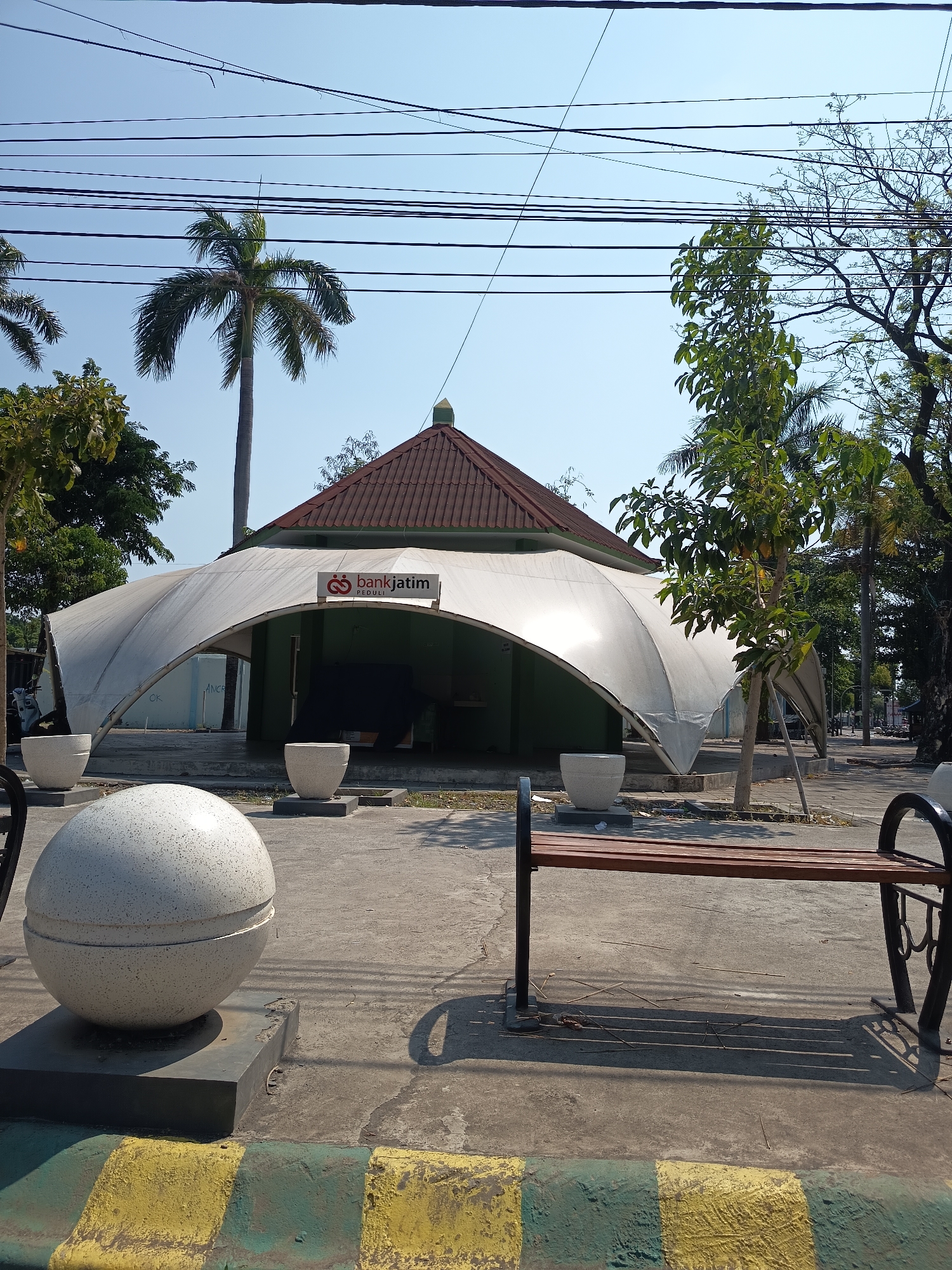 The image shows an outdoor scene in Kandangjati Kulon, Indonesia, featuring a unique building with a white tensile fabric roof supported by a green structure. The building has a prominent "bankjatim PEDULI" sign, indicating a branch of Bank Jatim. In front of the building, there are several large, light-colored planters and a decorative spherical sculpture. A wooden bench with black metal supports is visible in the foreground, casting shadows on the concrete ground. Palm trees and other foliage are present, suggesting a tropical environment. Overhead power lines crisscross the clear blue sky, and a pitched, tiled roof is visible above the white fabric structure. The overall impression is of a public space, possibly a bank branch or community center, designed with modern architectural elements.