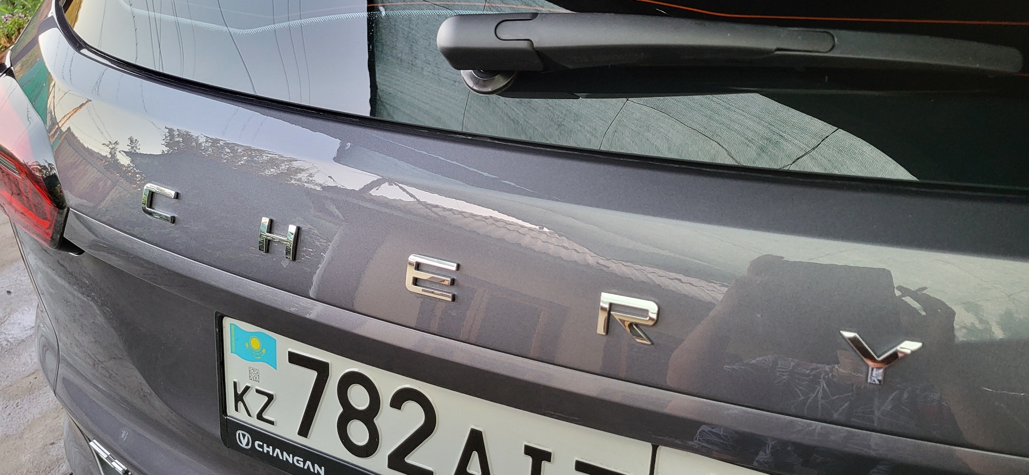 The image shows the rear of a grey car parked outdoors. The car has chrome lettering on the trunk that spells "CHERY". Below the lettering is a license plate with the text "KZ 782 AT" and the flag of Kazakhstan. The car is in a location that appears to be a residential area or parking lot, with a reflection of a house or building visible on the car's surface. The background includes a green mesh fence and some foliage. The license plate's "KZ" indicates Kazakhstan, and the city of Taldykorgan is a major city within Kazakhstan.