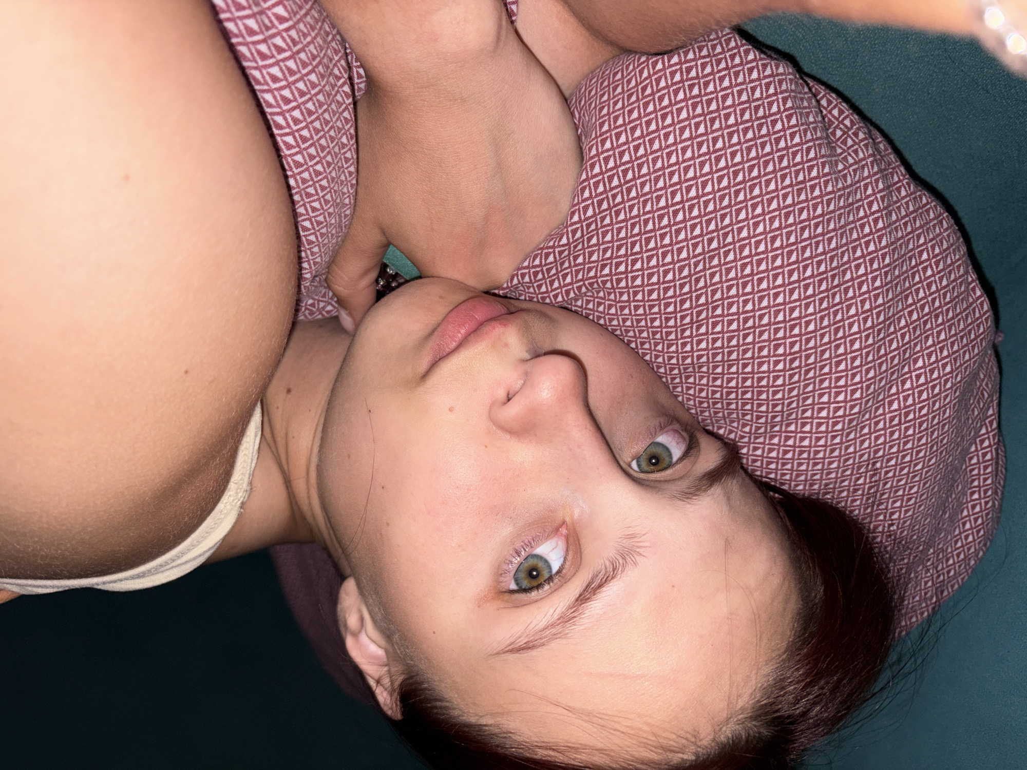 A woman is lying down, her head resting on a patterned pillow. She is facing the camera with her eyes open. A portion of a light-colored garment is visible at the top of the image. The background appears to be a dark green surface. No specific location in Прикамский, Russia is discernible from the image.