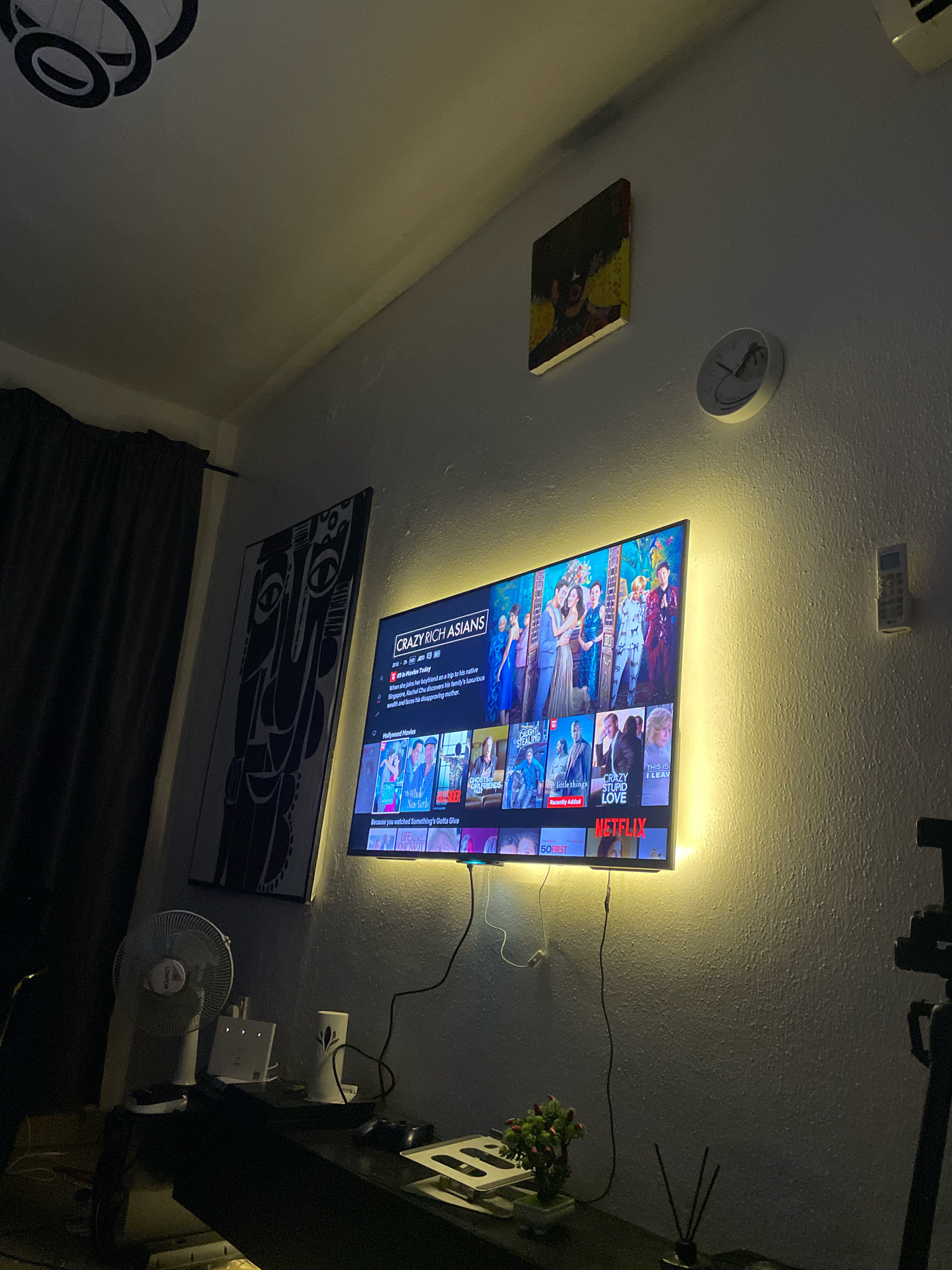 The image depicts a dimly lit room, likely a living room, with a large television screen mounted on a textured wall. The TV is displaying a Netflix interface, featuring movie posters including "Crazy Rich Asians." The television is surrounded by a warm, yellow LED light strip, creating an ambient glow. To the left of the TV, a tall, monochrome abstract art piece hangs on the wall. Further left, a dark curtain is visible. Above the TV, a small square artwork with yellow and black tones is displayed, and to its right, a round white clock with a minimalist design showing a palm tree motif.
Below the TV, a dark console table is present. On it sits a white electric fan, a white router, a white cylindrical object with a black design, and a small potted plant with red berries. A black game controller rests on the right side of the console. A tripod with a camera is positioned to the right of the TV, partially visible in the frame. The room appears to be in Ibadan, Nigeria, suggested by the presence of a "Crazy Rich Asians" movie poster which is a popular movie in Nigeria. The overall ambiance is one of relaxation and entertainment, with the lighting suggesting it's nighttime or the blinds are drawn.