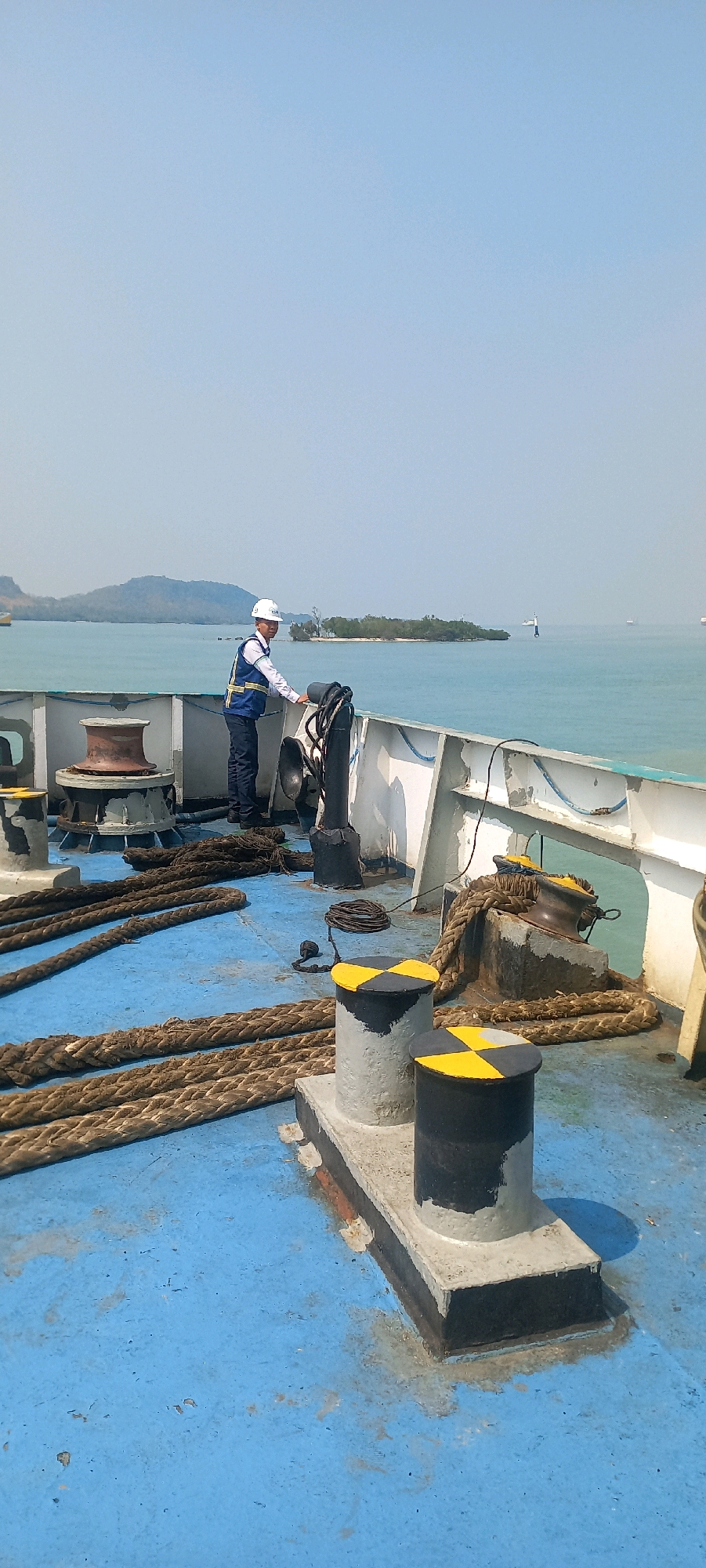 A full-frame, eye-level shot captures a deckhand in a white hard hat and a blue vest over a white shirt and dark pants standing on the blue deck of a ship. He is looking towards the water, his right hand resting on a metal railing. The deck is covered with thick, coiled ropes of varying thicknesses and a large anchor winch with a reddish-brown drum is visible to the left. Two grey bollards with black and yellow circular markings stand prominently in the foreground. In the background, the turquoise sea stretches to the horizon under a clear, pale blue sky. Distant landmasses, including a small island with trees and a larger, mountainous island, are visible on the left. A lighthouse and other vessels are scattered across the water in the distance. The scene is set in the waters near Bakauheni, Indonesia.