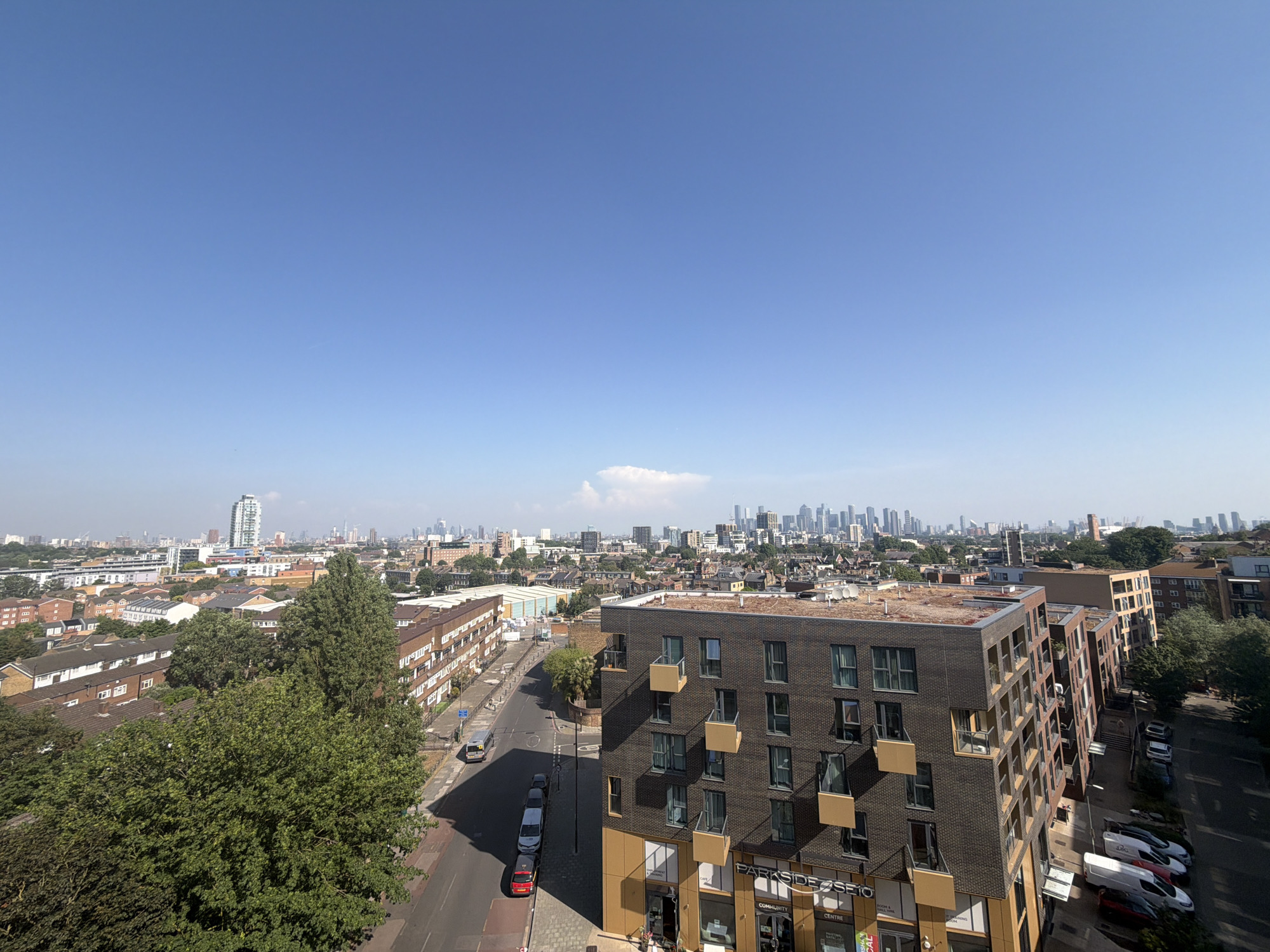 The image presents a cityscape in London, United Kingdom, under a clear blue sky. The perspective is elevated, offering a view that extends to the horizon. In the foreground is a modern apartment building with a dark brick facade and light-colored balcony recesses. A street runs alongside the building with parked cars. To the left, trees obscure some older residential buildings. In the distance, the skyline of central London is visible, with clusters of skyscrapers defining the urban landscape. A thin layer of haze hangs over the city.