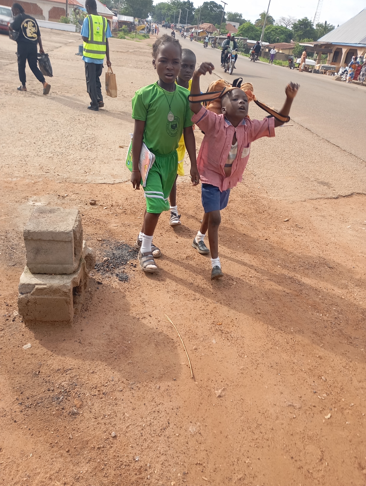 In the city of Shendam, Nigeria, on a dusty, unpaved road, two children are walking. A young boy in a red and white striped shirt and blue shorts is in the foreground, his arms raised, appearing to be in mid-stride. He is wearing a backpack with orange straps. To his left, a young girl in a green shirt and shorts is walking, holding a book. Behind them, other people and vehicles are visible on the road, suggesting a busy street. The environment is characterized by dry, reddish-brown soil, with some vegetation and buildings in the background. The lighting suggests it is daytime.
