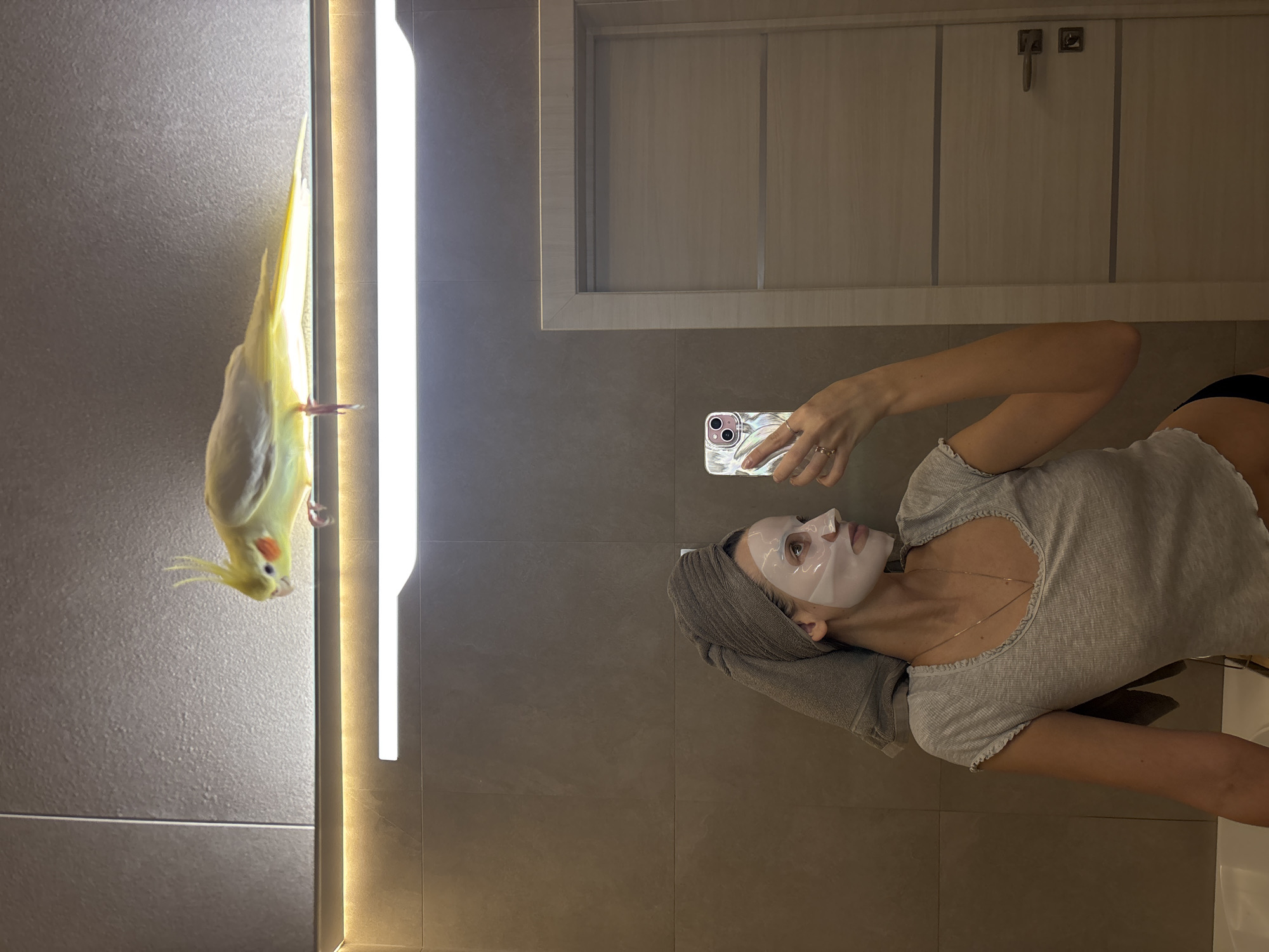 The image portrays a modern bathroom within a private residence in Krasnoyarsk, Russia. A young adult female is visible from the torso up, reflected in a mirror, actively taking a selfie with a smartphone. Her head is wrapped in a gray towel, and her face is covered with a white sheet mask. She wears a light gray ribbed short-sleeved top and a thin gold chain necklace. Her right hand, featuring two rings, holds an iridescent-cased smartphone. Perched on a horizontal LED light fixture directly above the mirror is a light yellow cockatiel with prominent orange cheek patches and an erect yellow crest. The bird faces left, partially turned towards the camera. The bathroom walls are clad in large, matte gray tiles. A light-colored wooden door with horizontal paneling and a modern silver handle is reflected to the right, and a white bathtub rim is partially visible in the bottom left of the reflection.