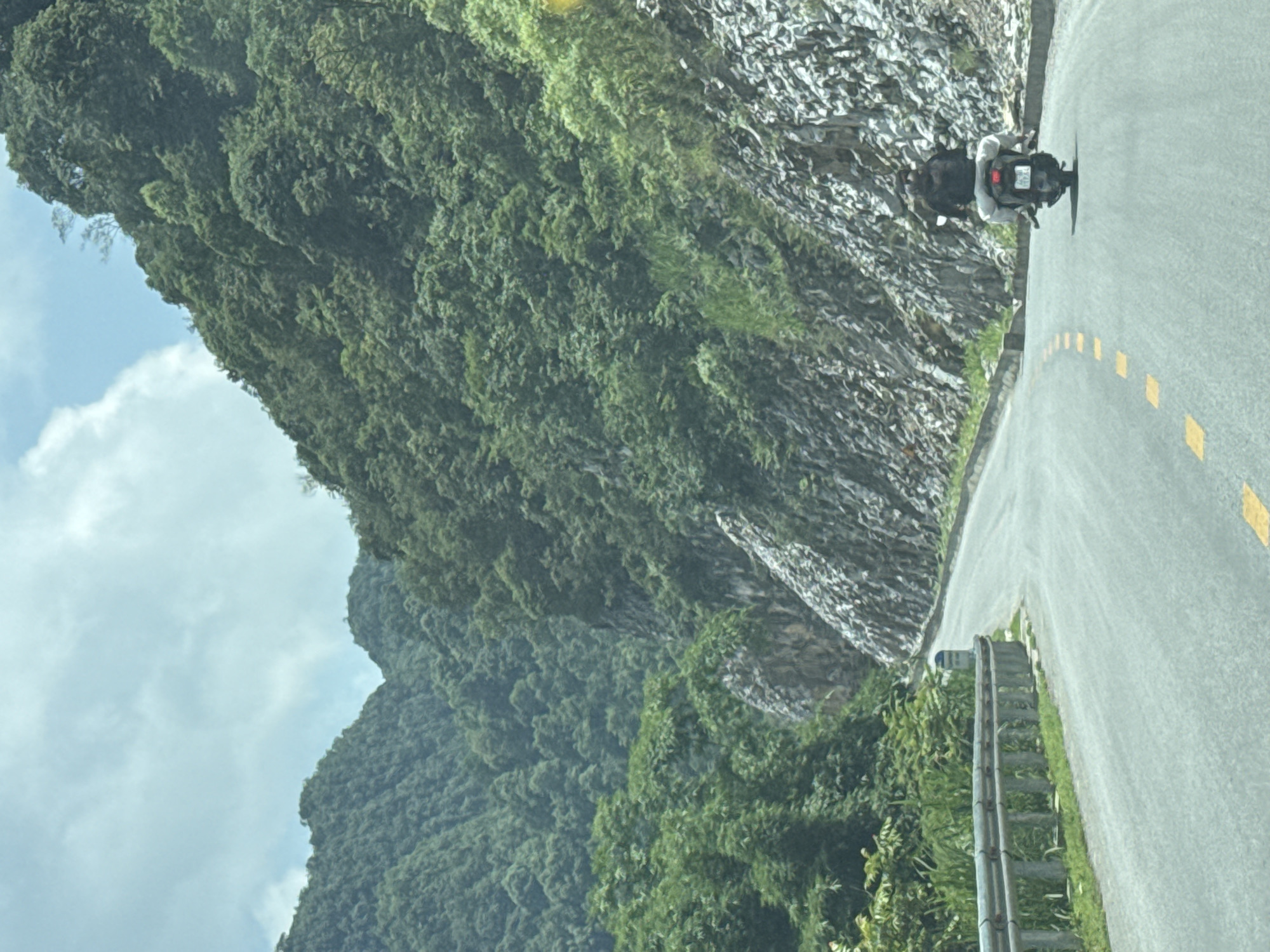 A motorcyclist travels along a paved road in Sủng Máng Commune, Vietnam. The road, marked with dashed yellow lines, curves along a steep, rocky hillside covered in lush green vegetation. A metal guardrail lines the edge of the road. Mountains and a cloudy sky are visible in the background.