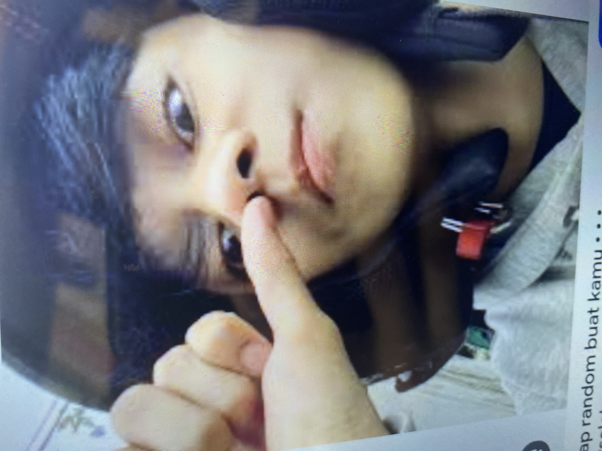 The image displays a close-up view of a young person wearing a dark helmet. Their right hand is raised, with the index finger extended and touching their nose. The person's eyes are wide, looking directly at the camera. The background is out of focus, but suggests an indoor setting with some text at the bottom. There is no identifiable location in the city of Ta', Indonesia.