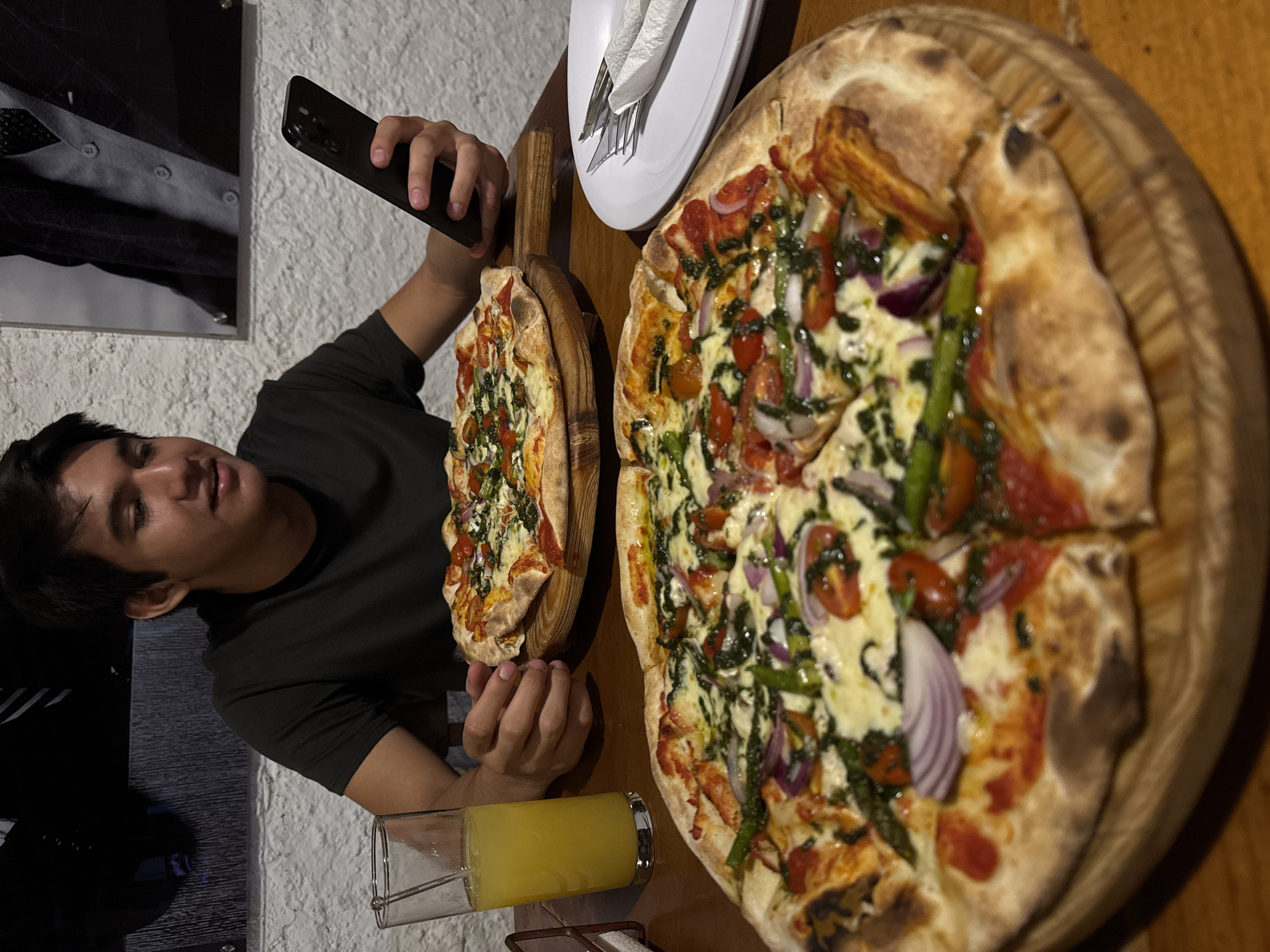 A young man in a dark green t-shirt is photographing two pizzas with his smartphone. The pizzas are on wooden serving platters. One pizza is smaller and appears to have a simple cheese and tomato topping. The larger pizza has toppings including tomatoes, red onion, spinach, and asparagus. A plate of utensils in a napkin is on the table between the pizzas. A glass of yellow liquid is in the foreground. The background features a partially visible framed print and a textured wall. The location is likely a restaurant in Chipilo de Francisco Javier Mina (Chipilo), Mexico.