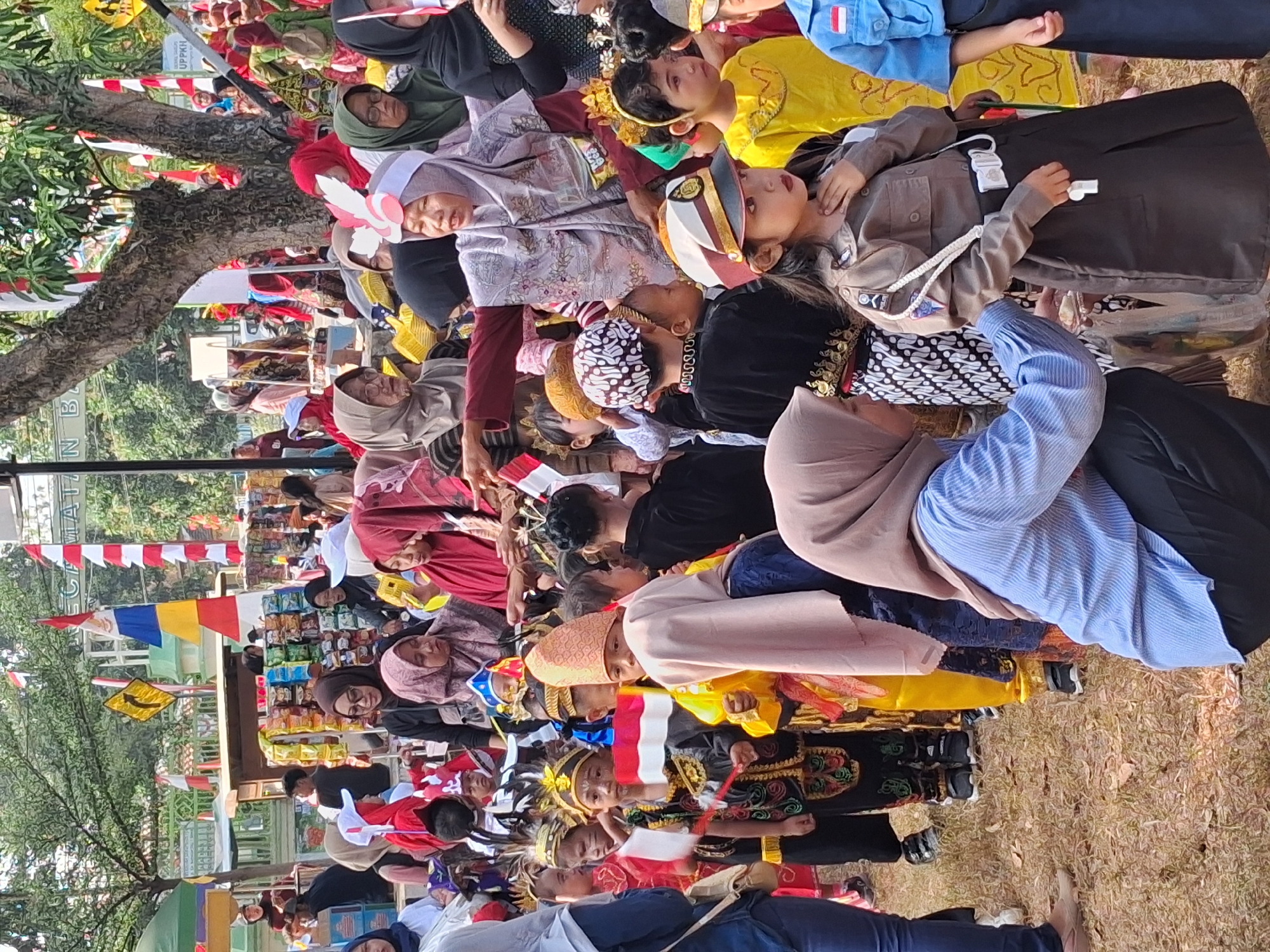 The image captures a cultural event, likely a children's parade or festival, taking place outdoors in the city of Balapulang, Indonesia. A crowd of people, predominantly women and children, are gathered. Many of the children are dressed in traditional Indonesian attire, including colorful outfits, headdresses, and face paint. Some children are holding small Indonesian flags, indicating a patriotic theme.
In the background, stalls are visible, possibly selling food or merchandise, decorated with Indonesian flags. Red and white striped flags are prominently displayed, along with other colorful banners. The environment appears to be a grassy area with trees providing shade. The overall atmosphere suggests a festive community gathering.
Specific details include:
* **People:** A diverse group of individuals, with a notable presence of children in elaborate costumes. Adults are also present, some supervising the children and others observing the event.
* **Objects:** Indonesian flags of various sizes, traditional clothing, headdresses, face paint, and what appear to be market stalls. One child is dressed in a police-like uniform.
* **Actions:** Children are standing in line, some holding flags, suggesting a parade or performance. Adults are interacting with the children and observing.
* **Environment:** Outdoor setting, likely a public space, with grass, trees, and market stalls.
The image portrays a vibrant cultural celebration rooted in Indonesian traditions, potentially commemorating a national holiday or local festival. The presence of children in traditional dress and flags strongly suggests a patriotic and communal event.