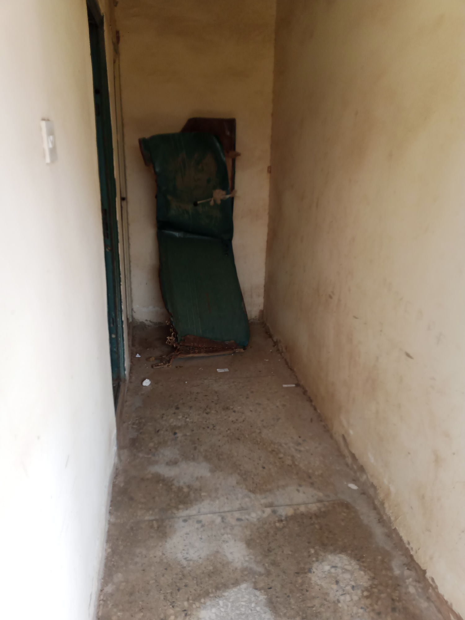 The image depicts a narrow, sparsely furnished hallway with light-colored walls that appear weathered and stained. On the left side, a white light switch is visible on the wall. The hallway opens into a slightly wider space where a dark green, padded object, possibly a deteriorated chair or mattress, is propped against a wall. The floor is a speckled concrete, showing signs of dampness and some debris. The overall environment suggests an abandoned or neglected interior space, likely within a building in Jalingo, Nigeria, given the context. There are no people present in the image.