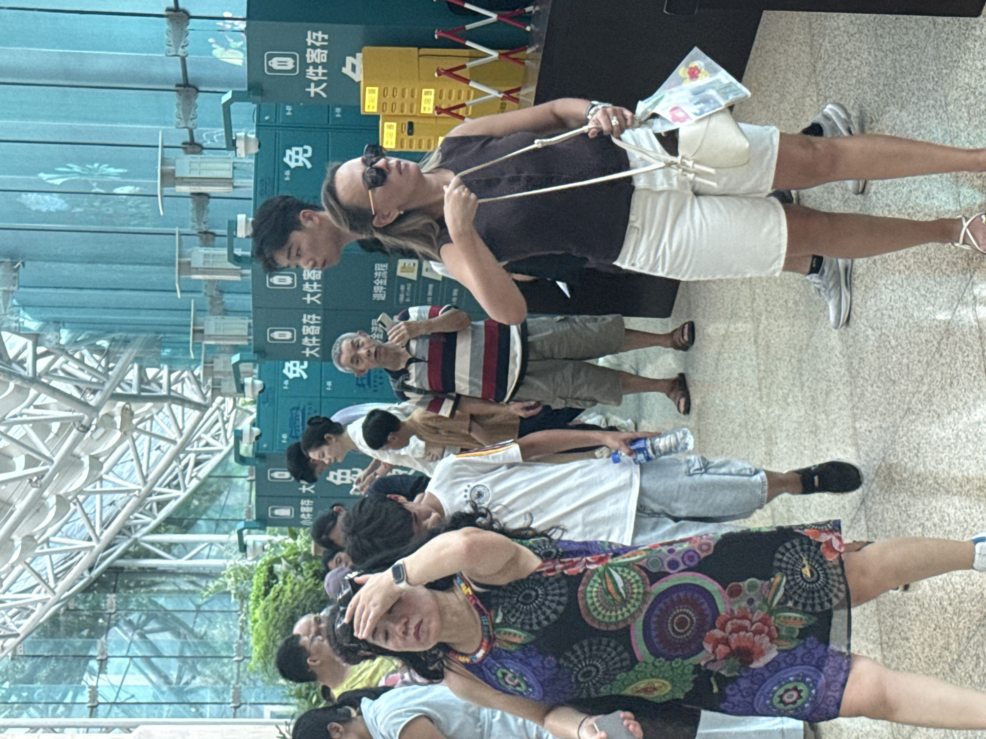 The image shows a crowded indoor scene, likely a transportation hub or public space, with a group of people visible. In the foreground, a woman with long dark hair and a colorful sleeveless dress is touching her forehead, appearing distressed or hot. To her right, a woman wearing sunglasses and white shorts is carrying a white handbag and a clear plastic bag with floral designs. Behind them, several other individuals are walking or standing, including a man in a striped shirt and shorts, and a young boy in a white t-shirt holding a bottle of water. In the background, there are rows of green lockers with Chinese characters indicating "大件寄存" (large item storage). The environment features a modern architectural design with a glass ceiling and metal beams. The scene is consistent with being in the city of Yuzhong District, China.