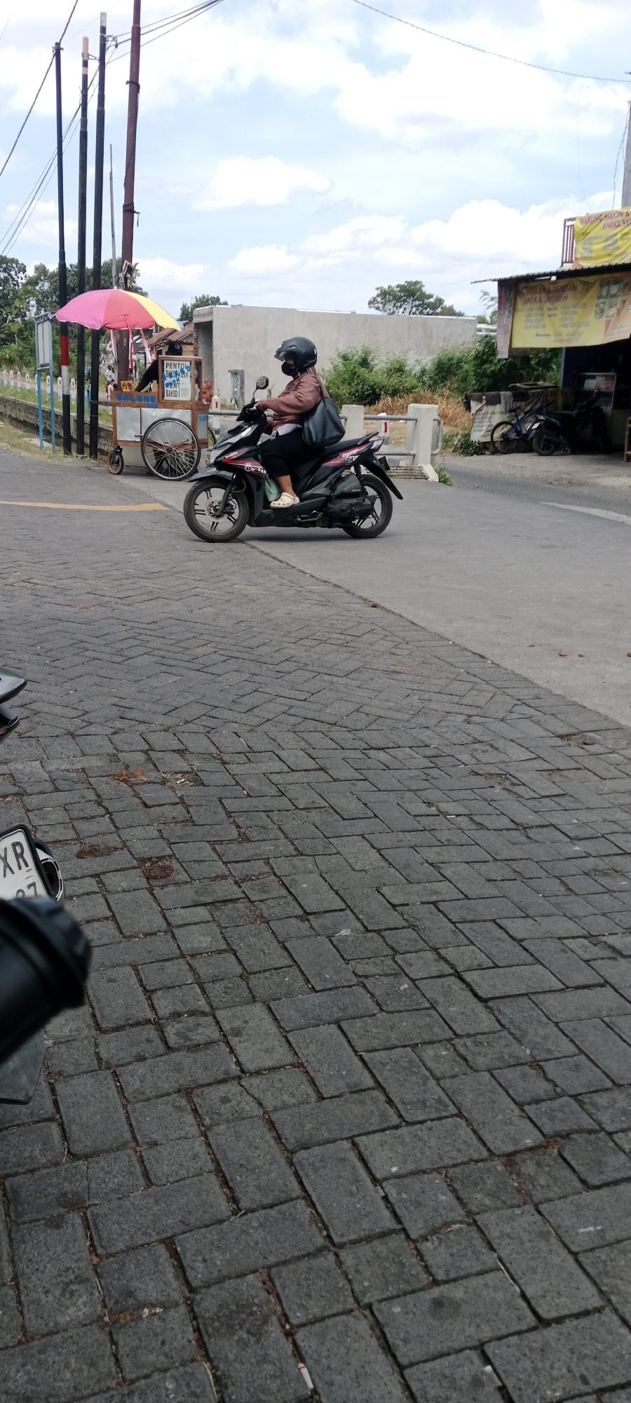 A medium shot captures a street scene in Maguwoharjo, Indonesia, focusing on a woman riding a black motorcycle. She wears a brown jacket, dark pants, and a black helmet, her face obscured by a mask. The motorcycle is in motion, turning slightly to the right. In the background, a street vendor's cart is visible, adorned with a colorful umbrella, and a sign displaying "PENTOL BAKSO". Further back, a building with yellow signage can be seen. The ground is paved with dark grey interlocking bricks. The sky is partly cloudy.