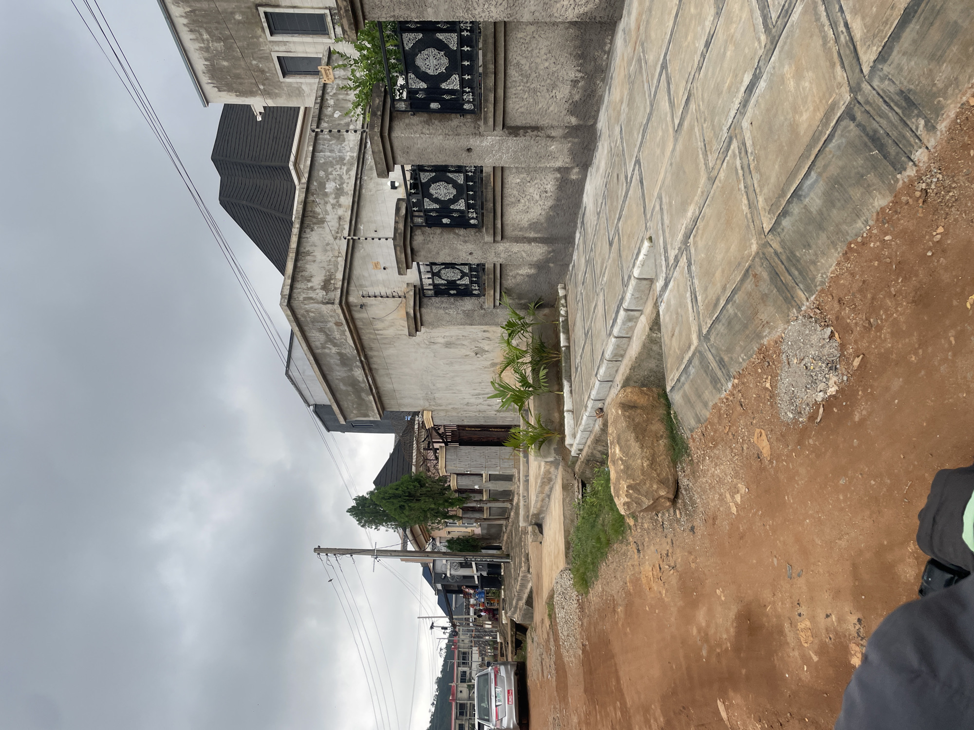 An overcast, cloudy sky looms above a street scene in Abuja, Nigeria. The foreground is dominated by a dirt road, rich in red soil, with a large, light-brown boulder resting on the side. Adjacent to the dirt road is a newly constructed concrete sidewalk, laid with square tiles and bordered by a concrete drainage channel.
The sidewalk leads to a row of buildings. The closest building on the right is a single-story structure with a gray, textured concrete facade. It features a black metal fence with ornate scrolling patterns supported by concrete pillars. Above the fence, an electric wire barrier is visible. To the left of this building, a similar structure stands, featuring a dark, sloped roof. Further down the street, other buildings can be seen, some with multi-story designs and dark roofs.
On the left side of the street, a silver sedan is parked. Beyond the buildings and houses, more structures and a scattering of trees line the horizon. A utility pole with several wires stretches across the scene. A few figures are visible in the middle distance, near one of the buildings, possibly interacting or walking. The overall impression is of a residential or commercial street in an area undergoing development.