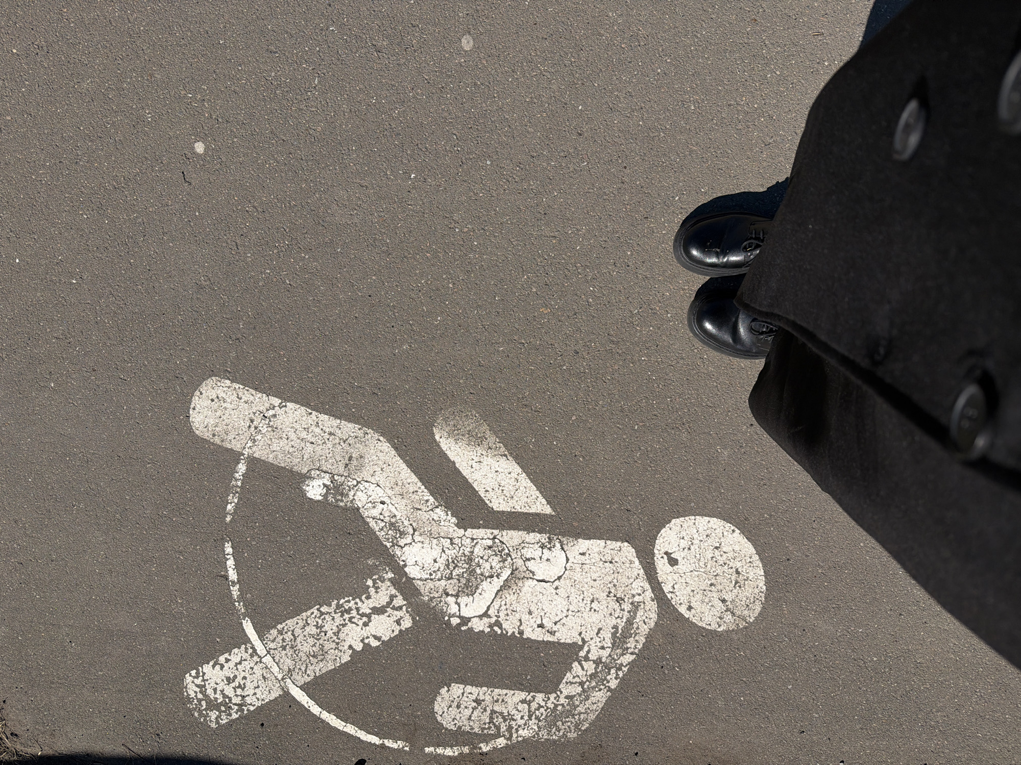 An eye-level outdoor shot depicts a gray asphalt surface with a faded white painted pedestrian crossing symbol indicating a person in motion. A person wearing black shoes and a long, dark coat with visible buttons stands partially in the frame, obscuring the upper right portion of the image. The asphalt is cracked and shows signs of wear. The location is in the city of Izhevsk, Russia.