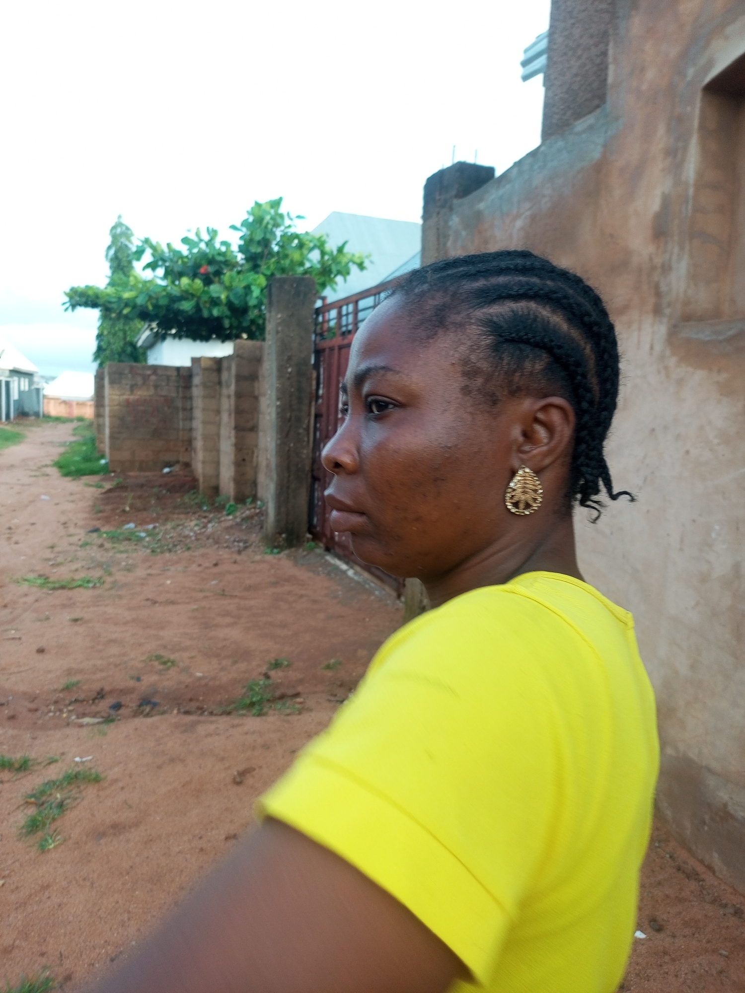 A medium shot shows a woman with braided hair and a bright yellow t-shirt facing to the left, with her head tilted slightly upward. She is wearing a teardrop-shaped gold earring in her right ear, which is adorned with a leaf-like pattern. The background features a sandy, dirt road with sparse green grass. To the left of the woman, a low brick wall is visible, behind which are some trees and buildings. To her right, a textured brown wall with a window opening is present, and further to the right is a brown metal gate. The sky is overcast and light-colored. The scene appears to be in an outdoor urban or semi-urban environment in Tempure, Nigeria.