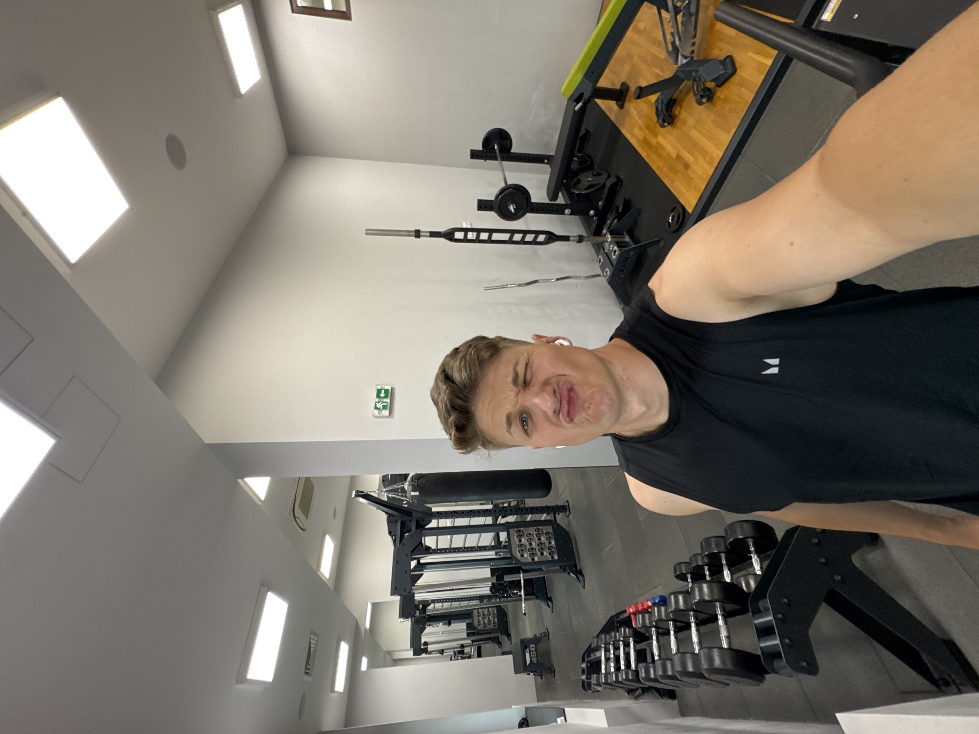 A man is taking a selfie in a gym. He is fair-skinned with short brown hair, wearing a black tank top, and has an earpiece in his right ear. His face is contorted in a grimace. The gym appears well-equipped with a weight bench and various barbells, dumbbells, and racks. A punching bag is suspended from a frame in the background. The gym is well-lit with rectangular lights. The location is Burgas, Bulgaria.