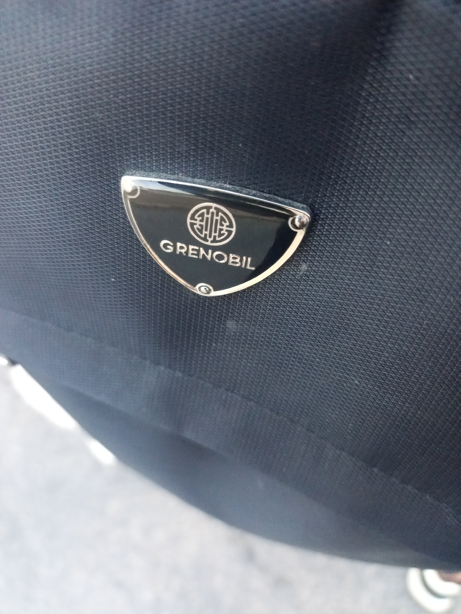A close-up shot focuses on a black textured fabric surface, likely a bag or luggage. A shield-shaped emblem is attached to the fabric, featuring the word "GRENOBIL" inscribed beneath a stylized circular logo. The emblem has a black interior with silver or chrome edging. The surrounding environment is out of focus, showing hints of white and gray tones suggesting a possibly outdoor setting. There are no discernible people or activities in the frame. The location is not identifiable as Juan Griego, Venezuela based on the available image.